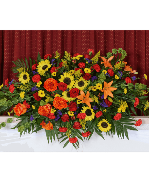 Radiant Tribute - A vibrant casket spray full of life and color—featuring sunflowers, orange lilies, red roses, and bold greenery. A radiant tribute that celebrates a beautiful soul with warmth, energy, and love.