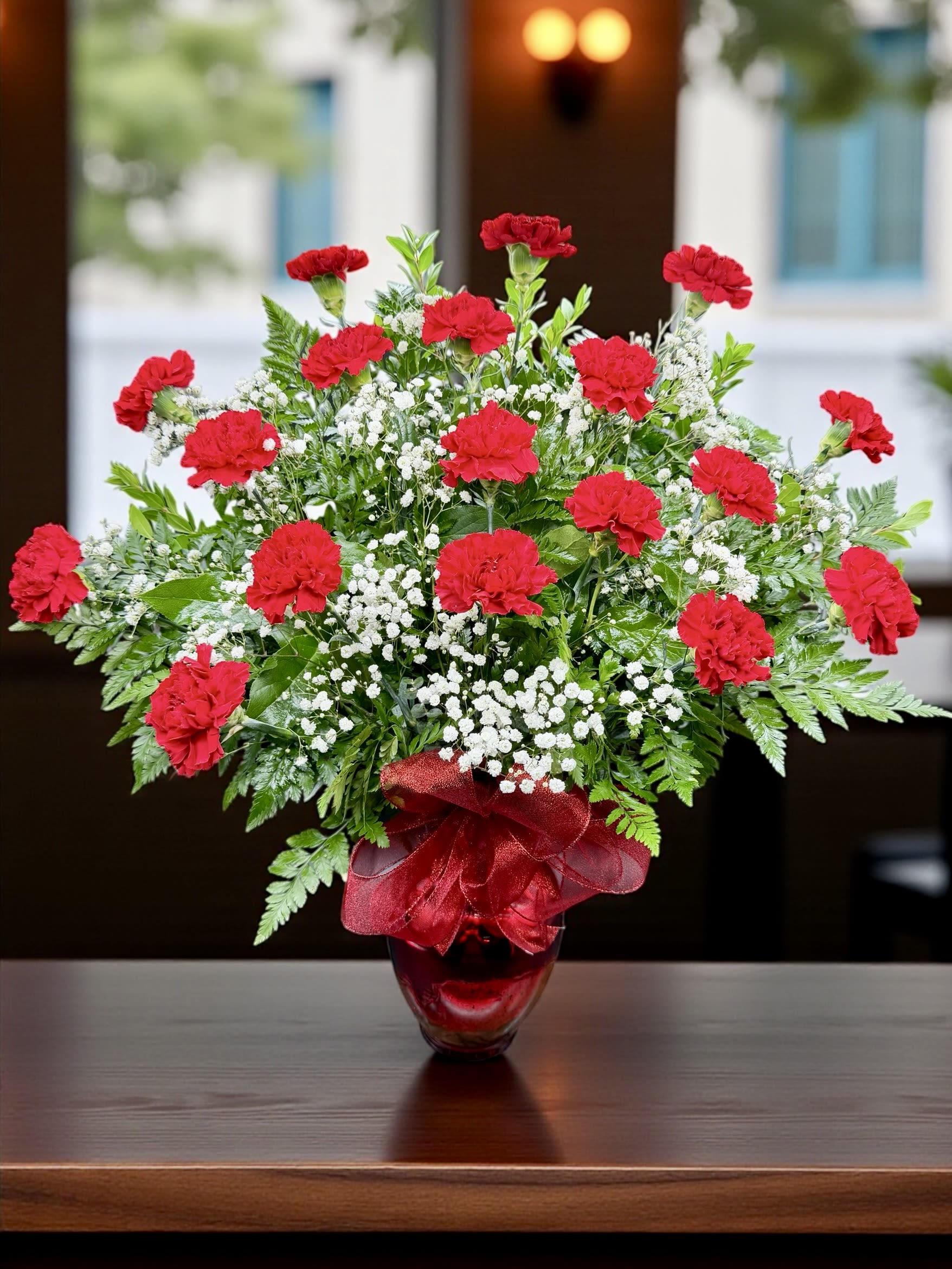 Red carnations and white baby's breath in a red glass vase with a ribbon