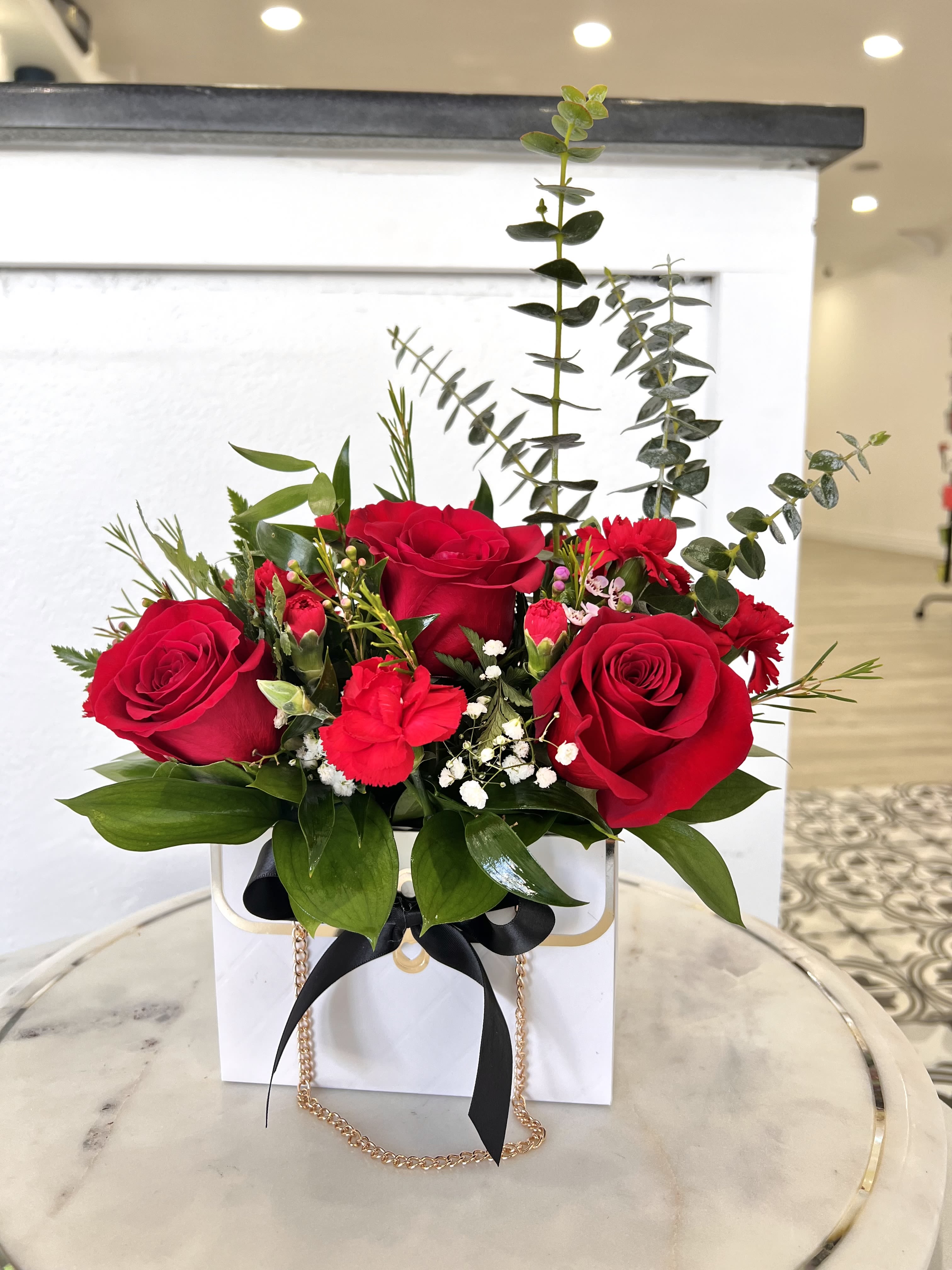 Red roses arranged in a white gift box with greenery and a black ribbon