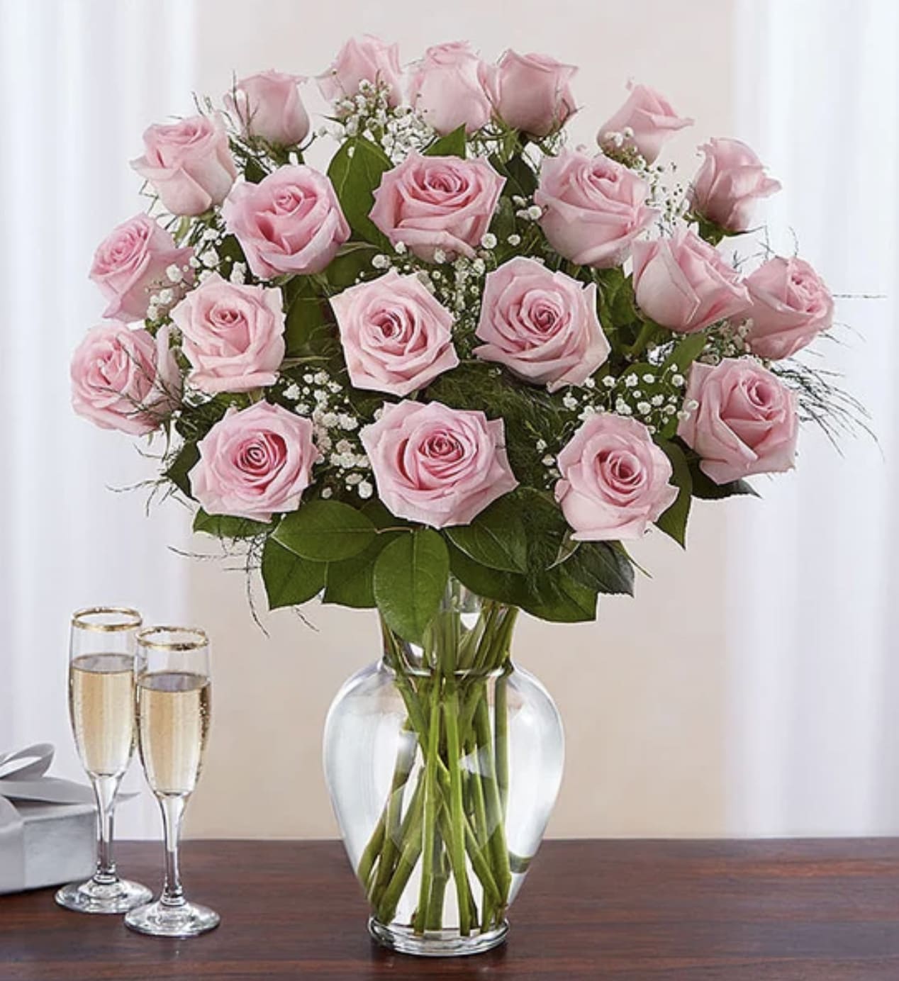 Tall arrangement of light pink roses with white filler flowers in a clear glass vase on a table