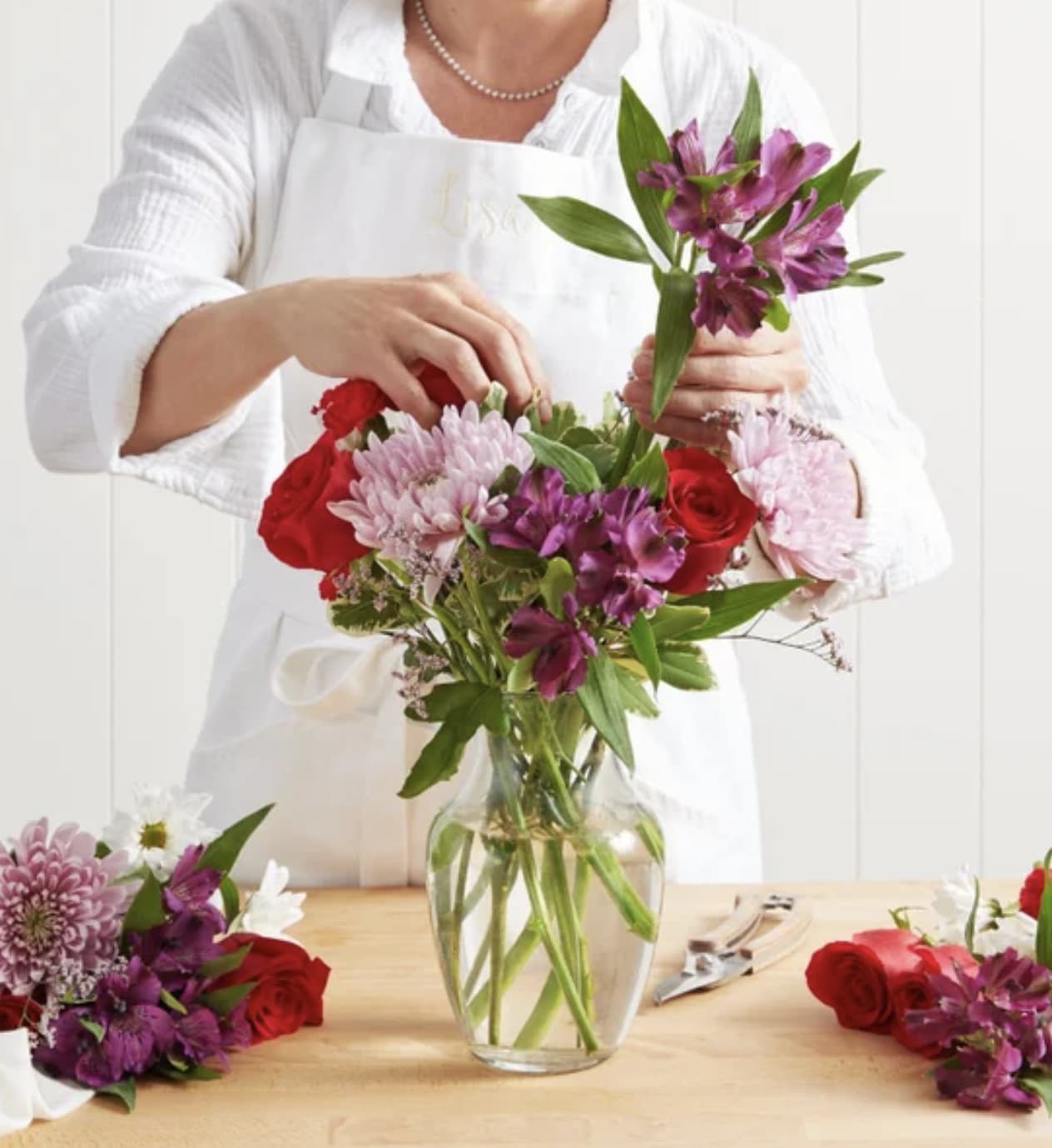 Person arranging a bouquet of red, pink, and purple flowers in a glass vase