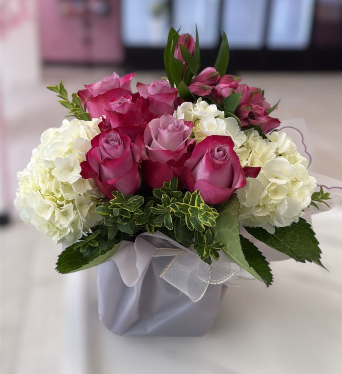 Compact arrangement of pink and lavender roses with white hydrangeas in a square vase with ribbon.