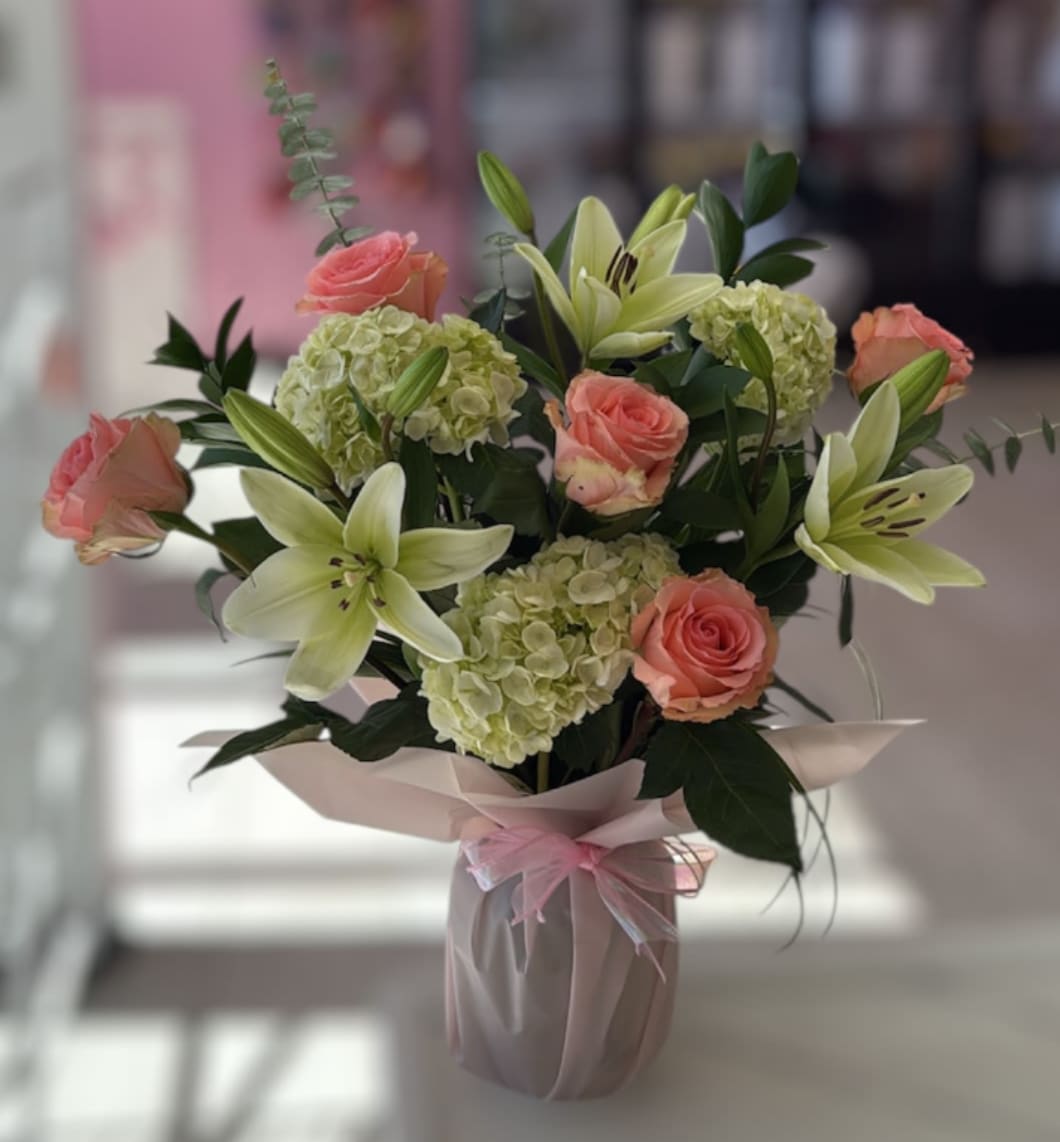 Vase arrangement of peach roses, green hydrangeas, and white lilies wrapped with pink paper.