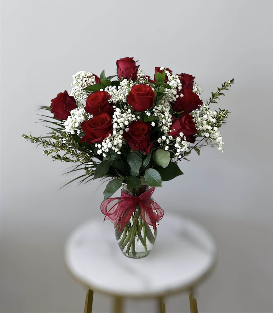 Clear glass vase of red roses with white filler flowers and a red bow.