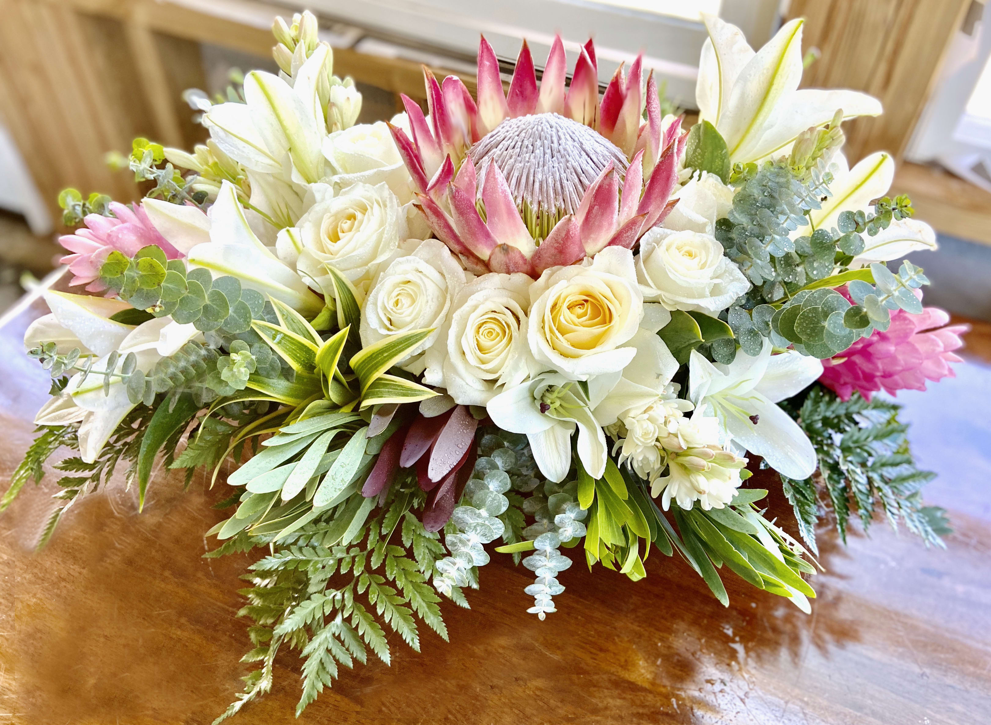 Low arrangement of white roses, lilies, and a pink protea with lush foliage on a wooden table