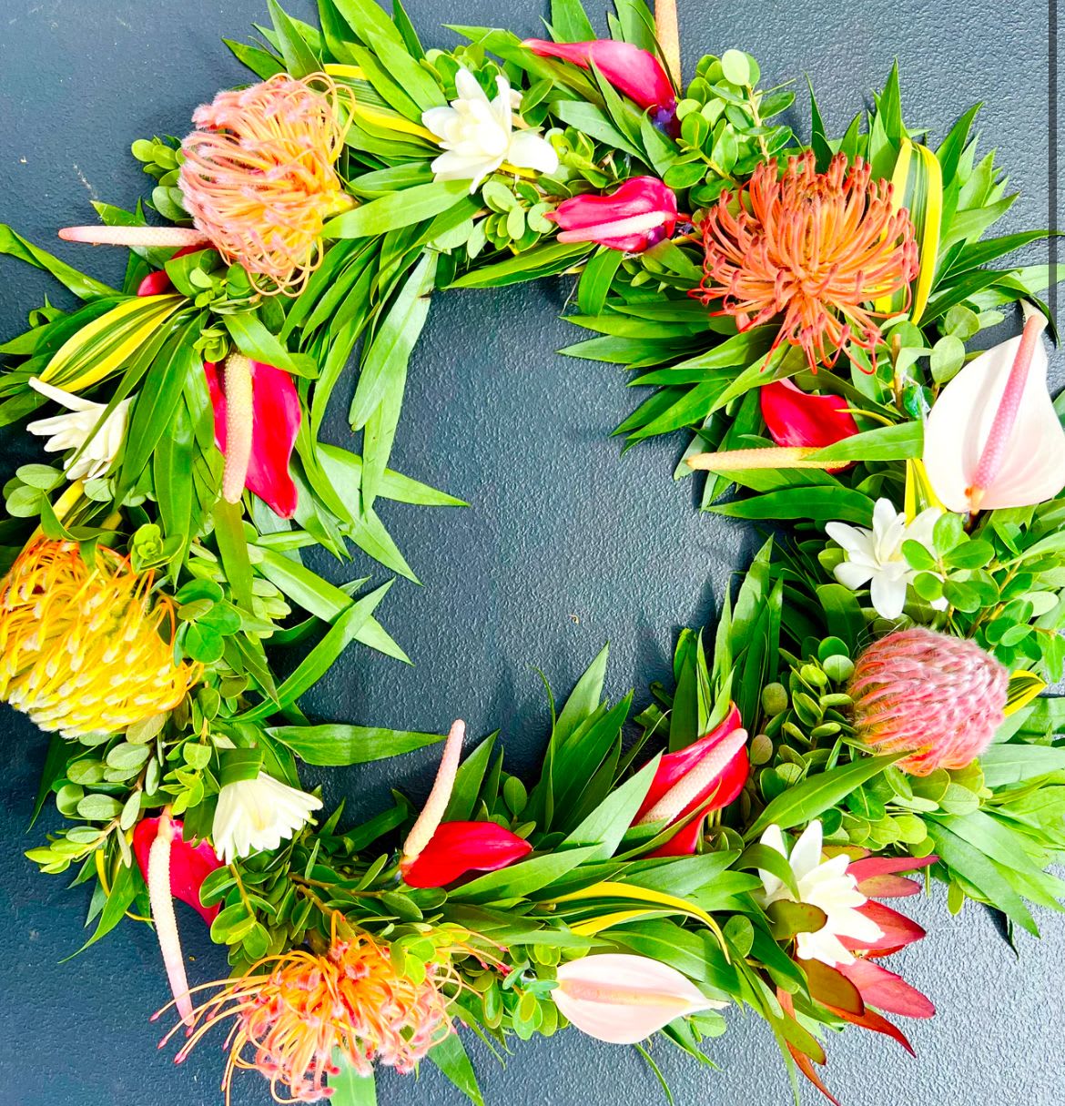 Tropical wreath of green foliage with protea and red, yellow, and white flowers on a dark surface
