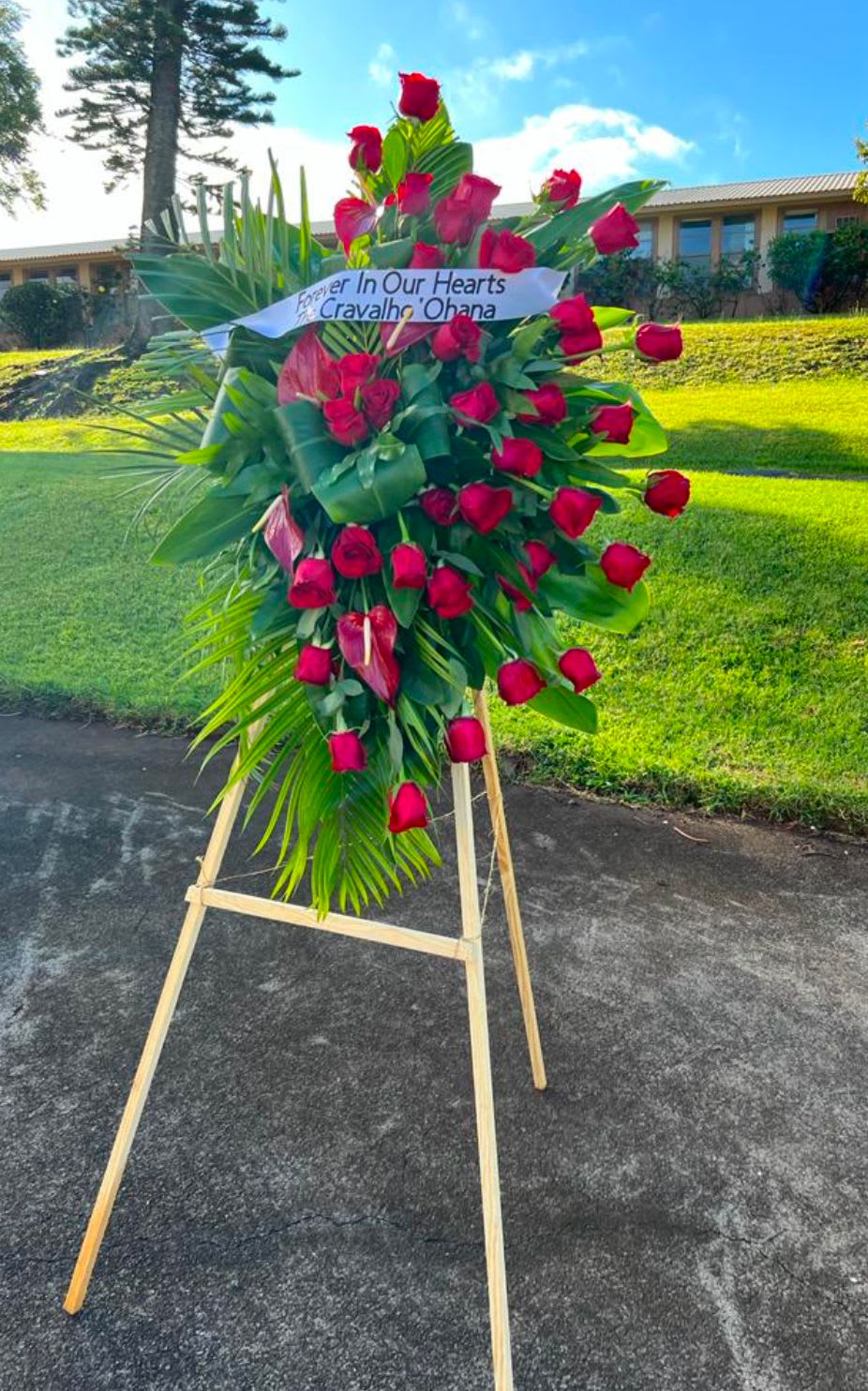 Standing spray of red roses and tropical blooms on an easel with a memorial banner.
