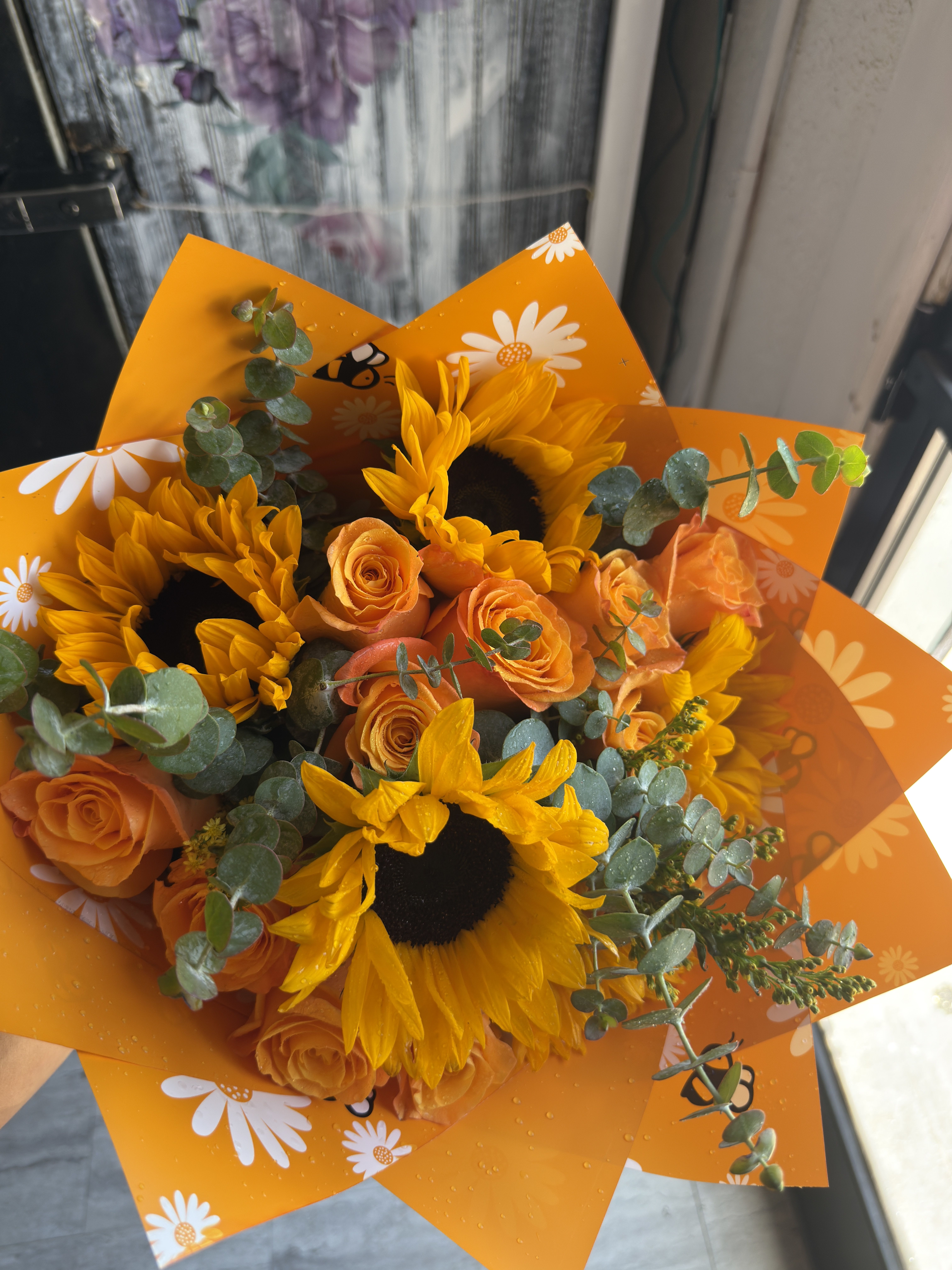 Honeybee harvest - A sunny mix of bright yellow sunflowers and warm orange roses makes this flower arrangement a cheerful pick for a birthday bouquet or a just-because celebration. The Honeybee harvest arrangement reads joyful and seasonal-perfect for brightening a kitchen table or making the person you're thinking of feel noticed and celebrated.  Tightly clustered sunflowers and orange roses are softened by airy eucalyptus and touches of pink and green, giving a full, hand-tied shape with pleasing texture and contrast. The stems are wrapped in a vivid orange decorative floral wrap printed with white daisies, so the presentation arrives ready to display and keeps that playful, upbeat look.  Choose this arrangement when you want someone to smile-ideal for friends, coworkers, or anyone who loves bold, sunny color. Vase-ready and arranged for immediate impact; light trimming of stems and fresh water will keep the blooms vibrant as they settle into their new home.