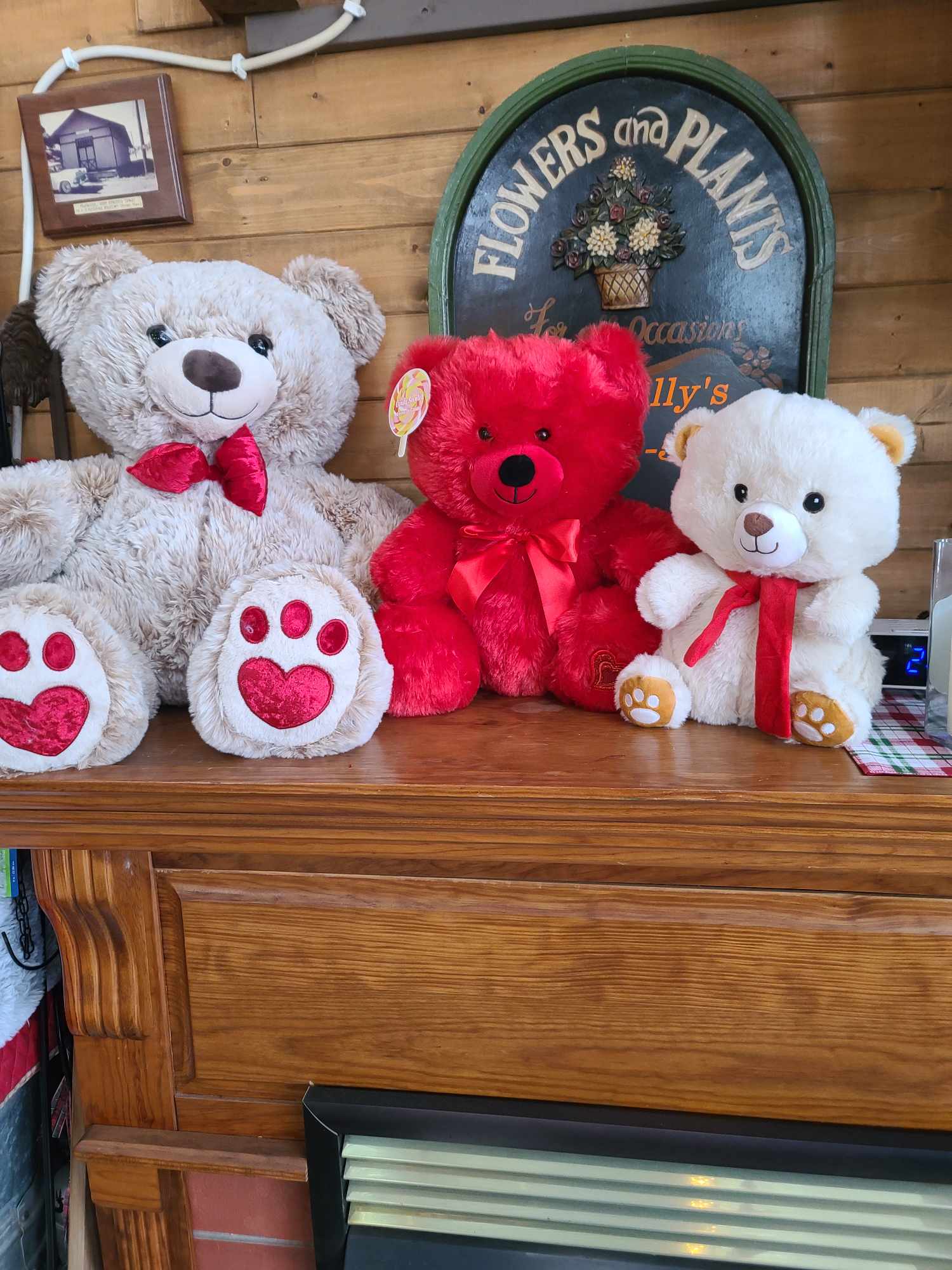 Three teddy bears with red ribbons sit on a wooden mantel.