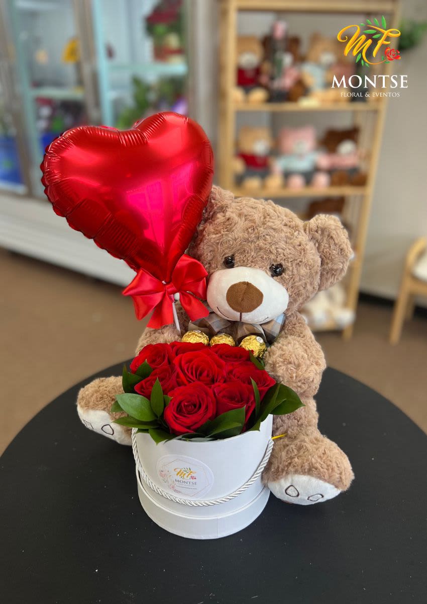 Teddy bear holding a red heart balloon beside red roses in a white box