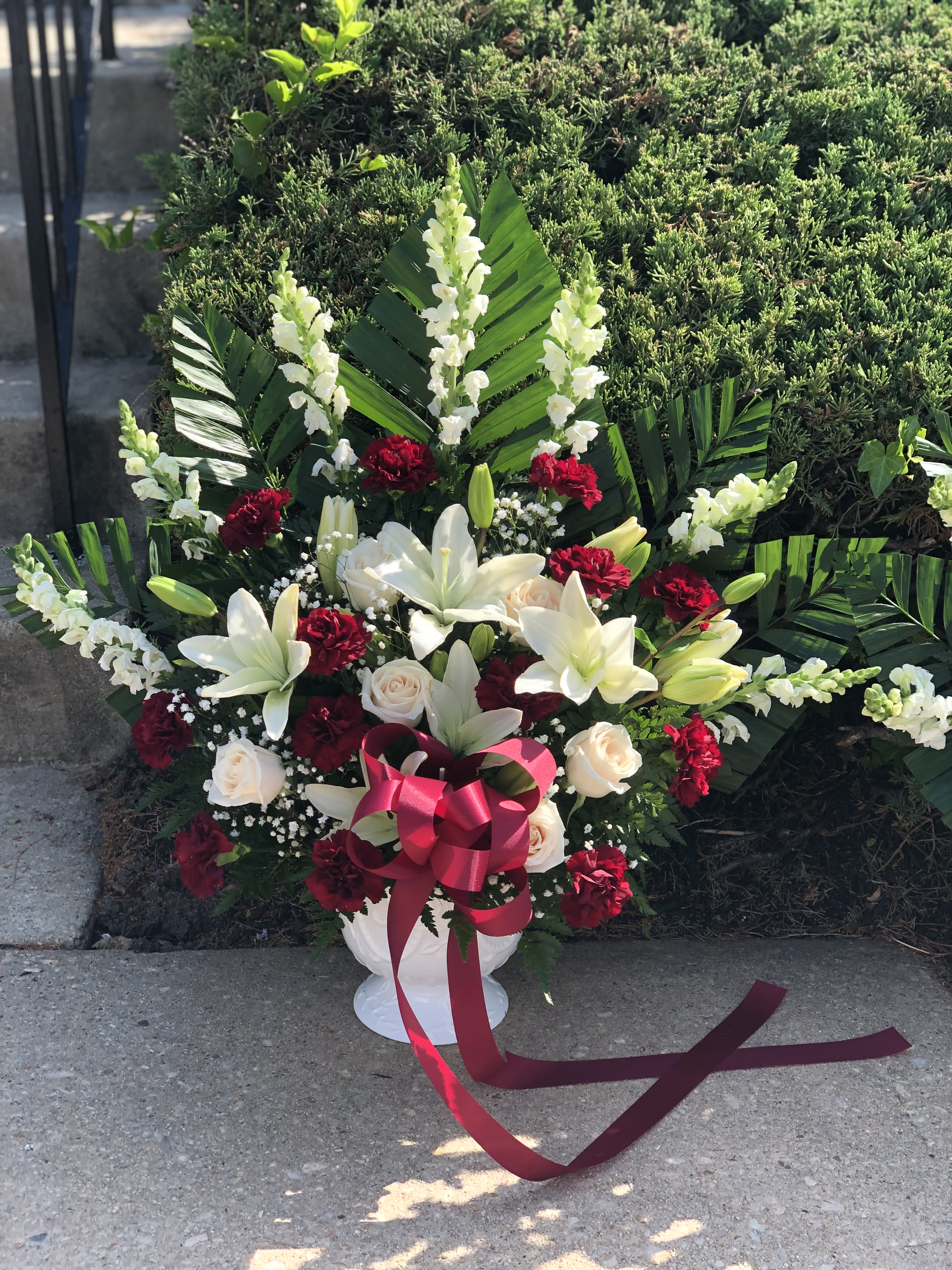 White lilies and red carnations arranged in a white urn with a pink ribbon