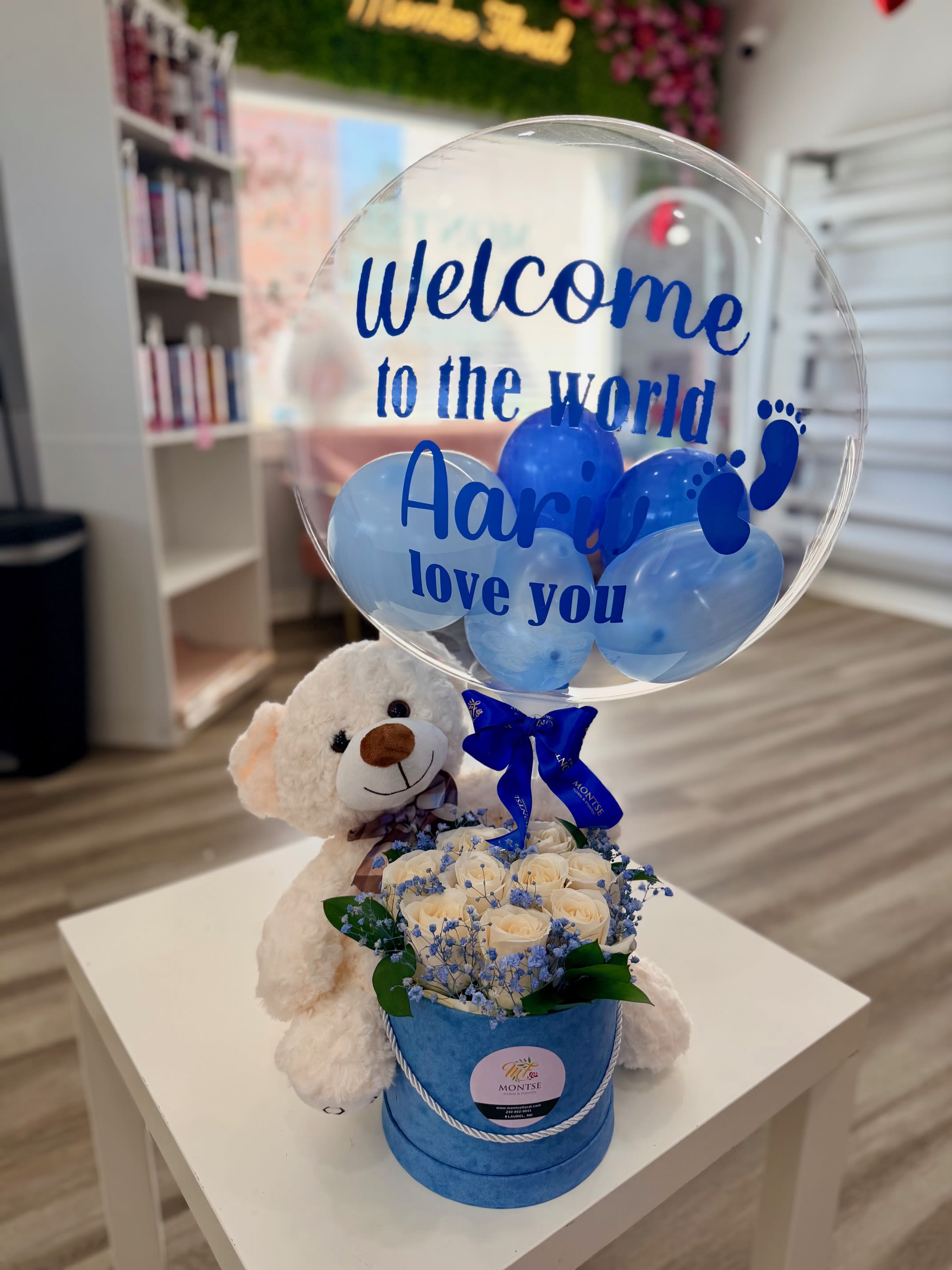 Cream teddy bear with box of white roses and clear balloon with blue text and balloons.