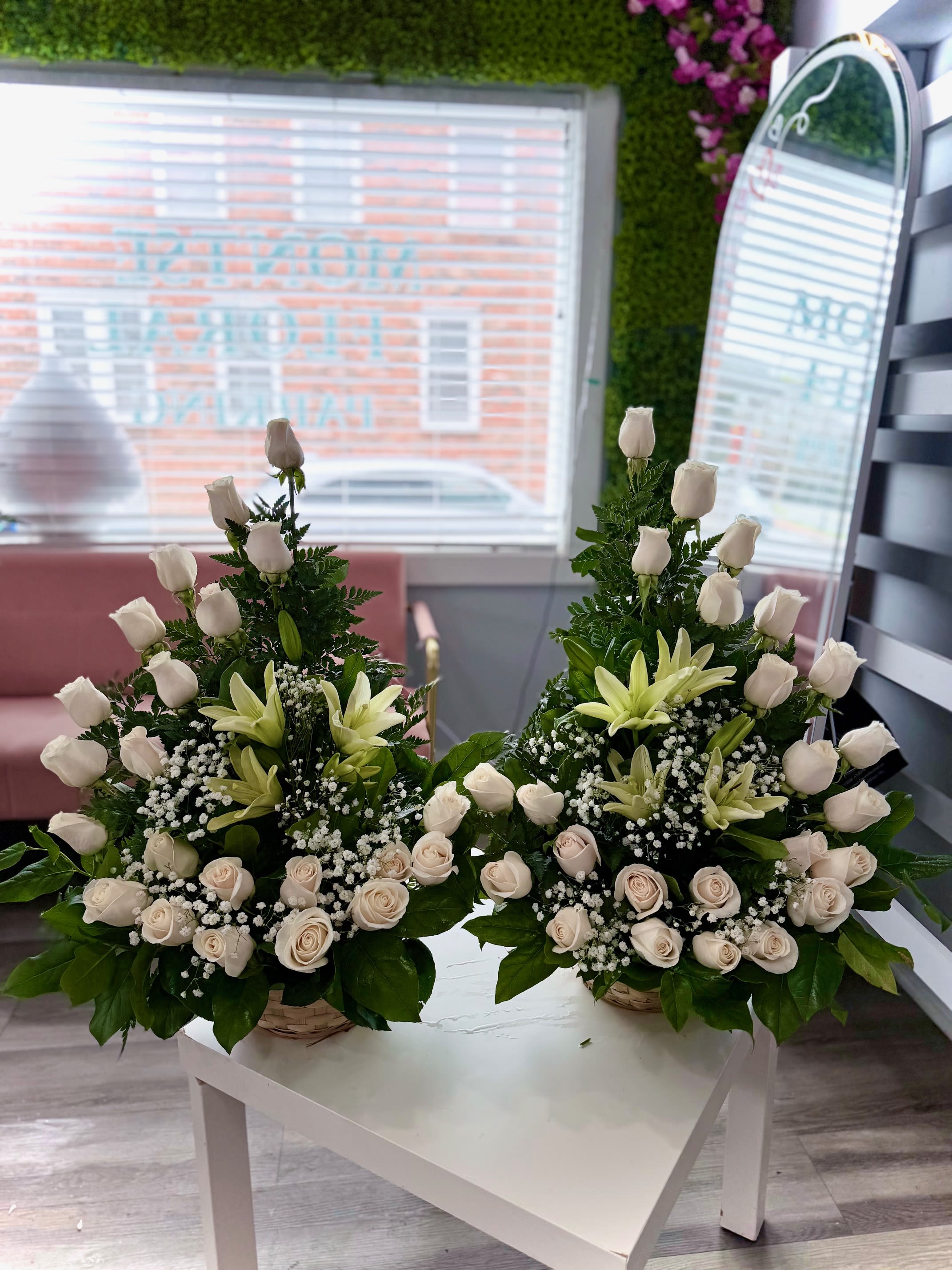 Pair of white rose and lily arrangements with baby's breath in woven baskets on a white table.