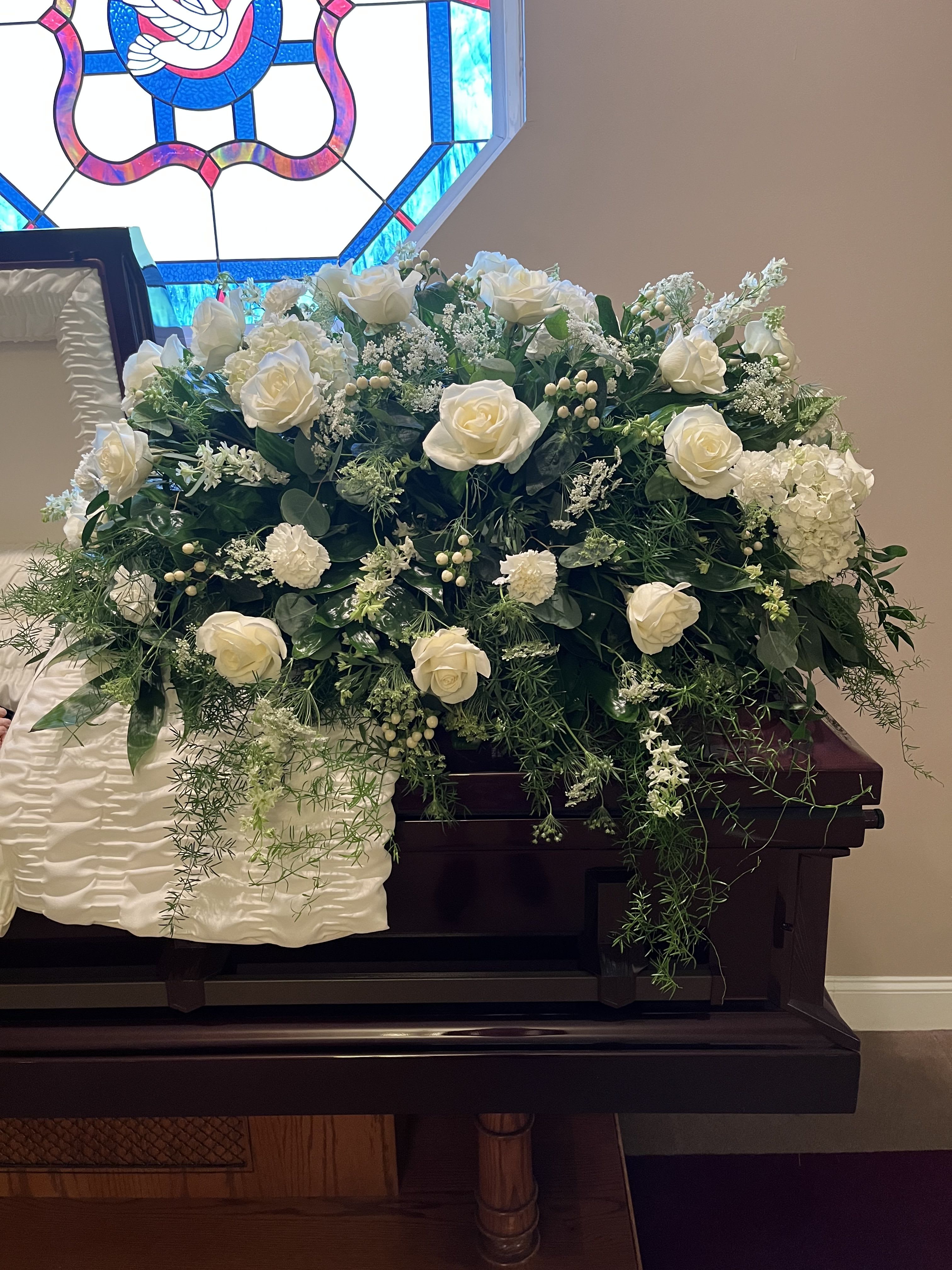 White rose arrangement on a dark wooden casket with trailing greenery