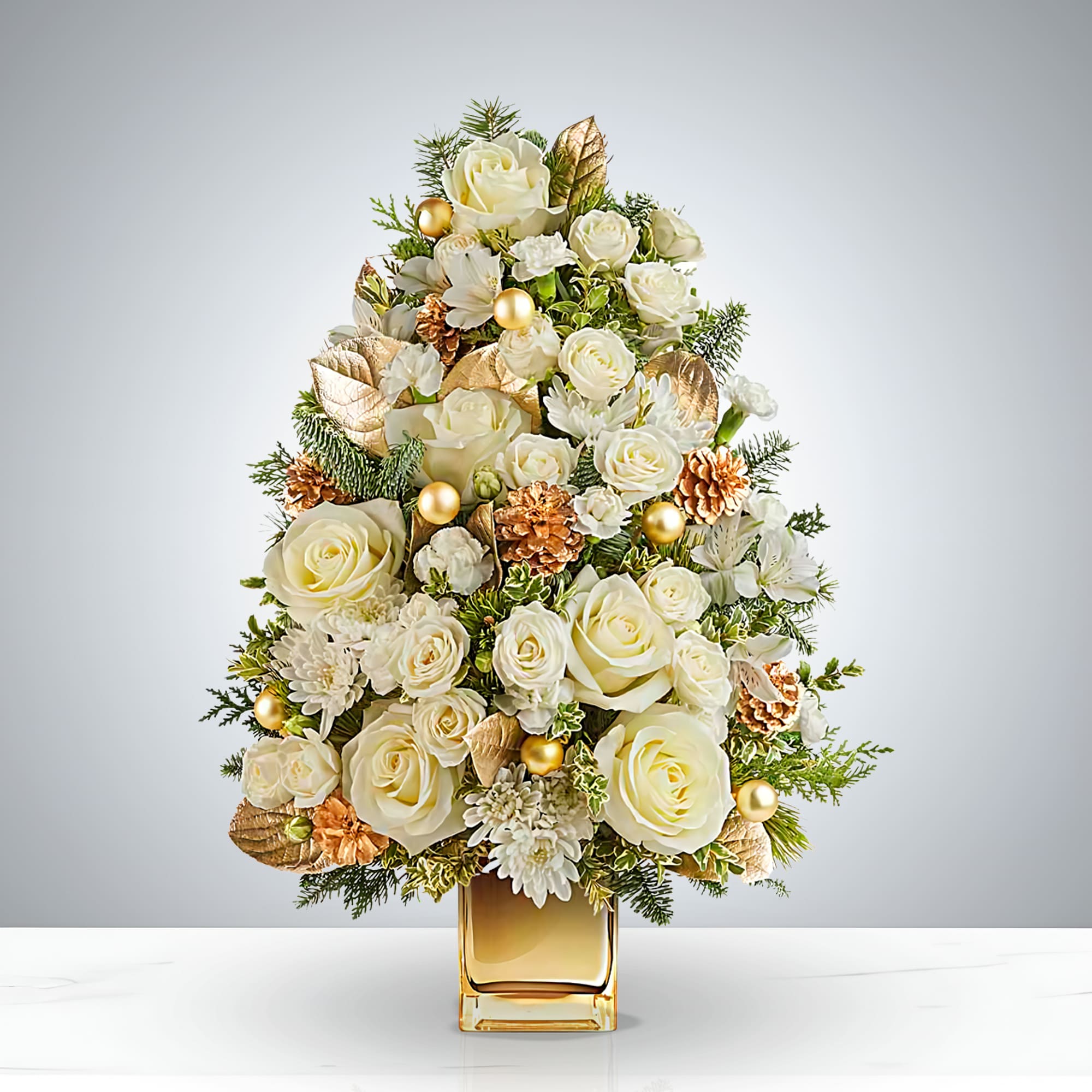 White Noel Luxe Tree - Celebrate the season in style with the White Noël Luxe Tree, a handcrafted Christmas floral arrangement that radiates elegance. Featuring creamy white roses, soft chrysanthemums, and lush pine greenery, this festive design is accented with shimmering gold pinecones, leaves, and ornaments.  Presented in a golden square vase, the White Noël Luxe Tree makes a stunning holiday centerpiece, perfect for home décor, office displays, or as a refined Christmas gift. A true symbol of luxury and cheer, it brings timeless beauty to every celebration.  Orientation: One-Sided