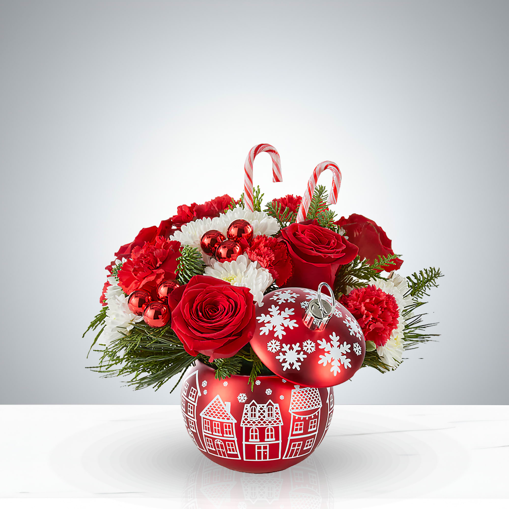 Candy Cane Christmas - Bring the magic of Christmas into your home with Candy Cane Christmas, a charming holiday floral arrangement displayed in a red ornament‑shaped vase adorned with snowy village illustrations. This cheerful centerpiece features radiant red roses, festive carnations, snowy white chrysanthemums, and fresh pine sprigs, accented with shiny red ornaments and playful candy canes. Perfect for dining tables, entryways, or as a thoughtful seasonal gift, it captures the warmth and joy of the holidays in every detail.