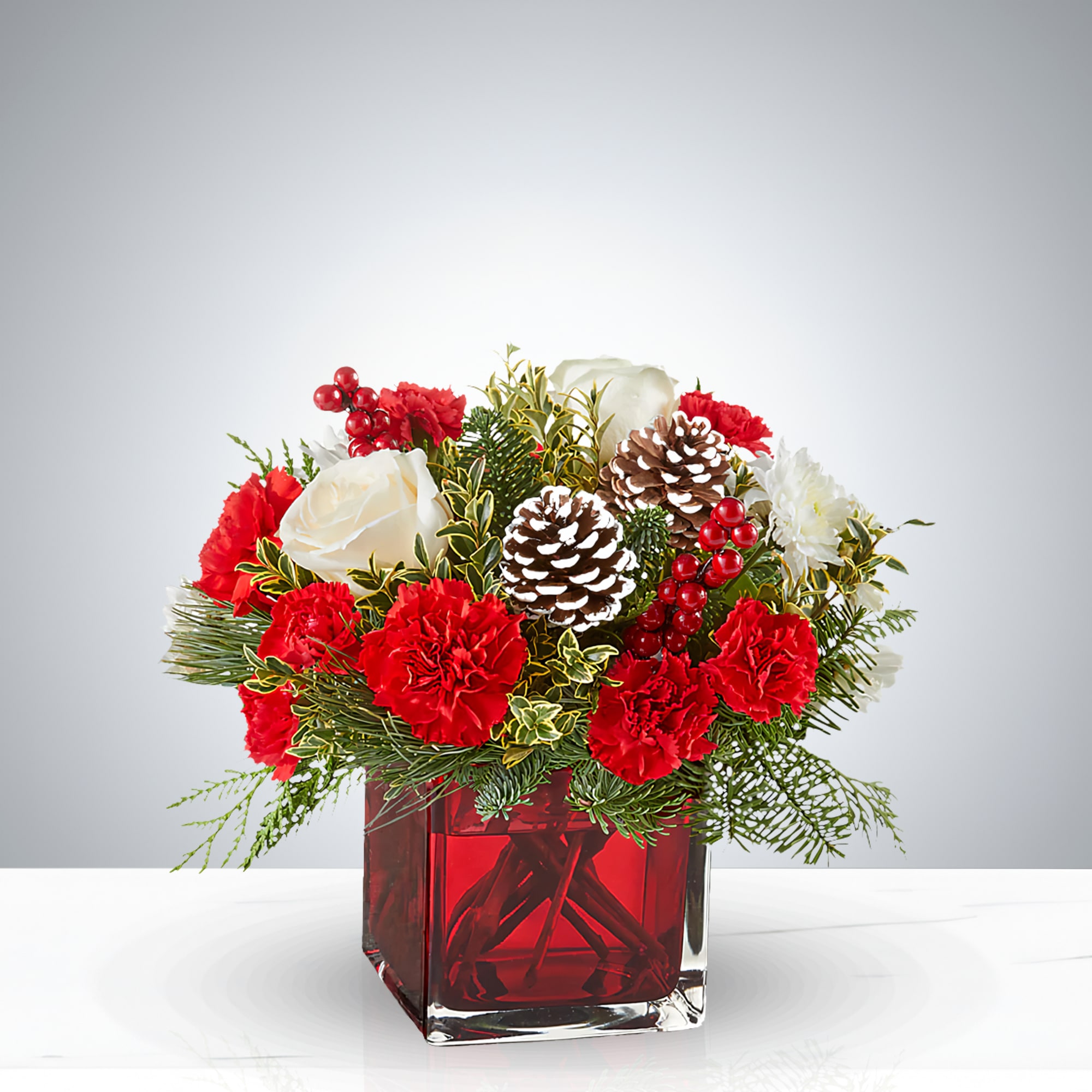 Red Christmas Elegance - Celebrate the magic of the season with this luxurious Christmas floral centerpiece, artfully arranged in a sleek red glass vase. Featuring velvety red carnations, creamy white roses, frost-tipped pinecones, festive red berries, and lush evergreen sprigs, this bouquet captures the warmth and elegance of the holidays. Perfect for holiday tables, Christmas gifts, or seasonal home décor, this arrangement blends classic Christmas colors with natural textures for a timeless look. Whether you're hosting a festive dinner or sending cheer to loved ones, this handcrafted design brings joy and sophistication to any space.
