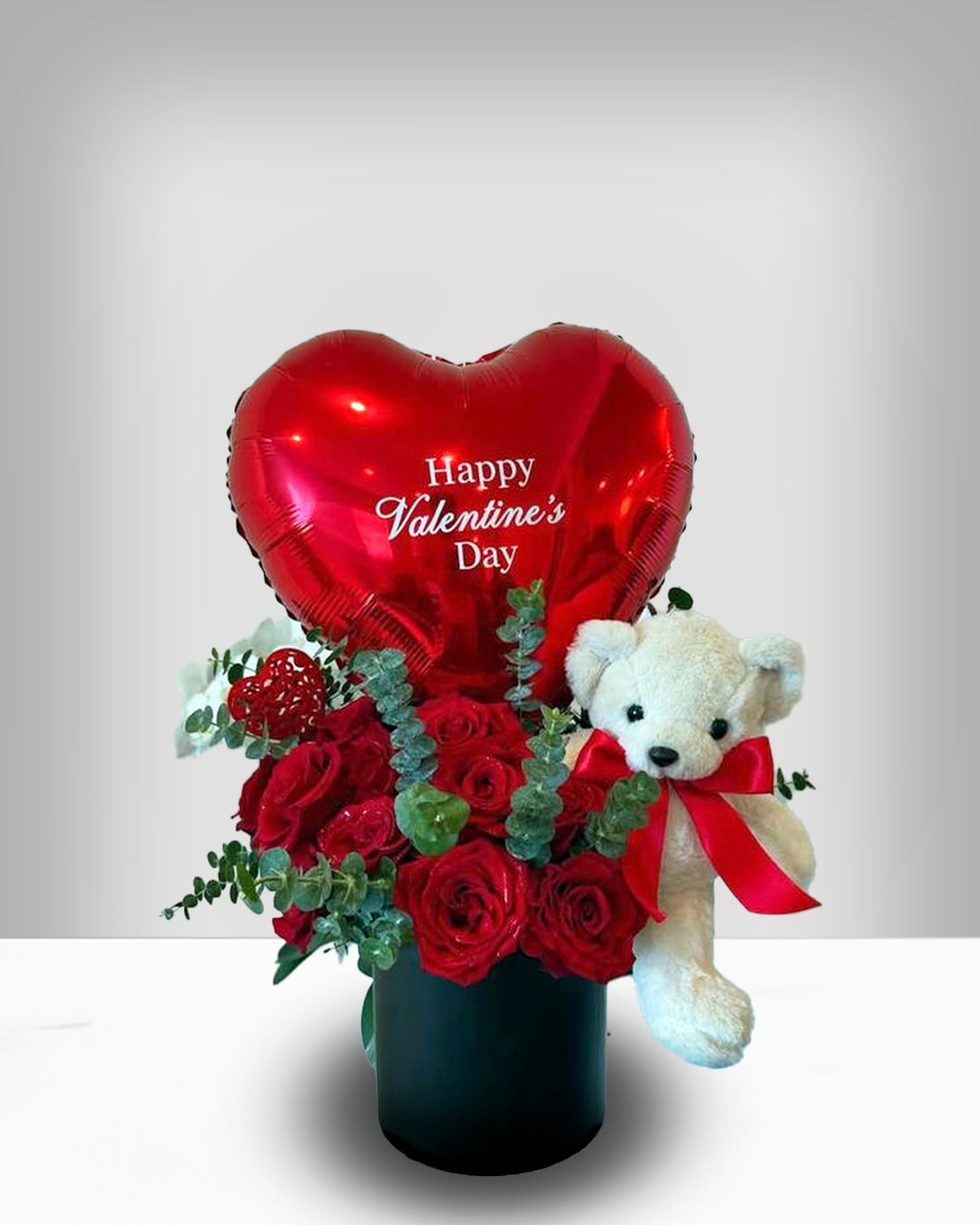 Luxury Love - A romantic arrangement featuring a heart‑shaped “Happy Valentine’s Day” balloon, fresh red roses with greenery, and a plush white teddy bear with a red ribbon. Presented in a sleek black vase, it’s the perfec gift for expressing love and affection.