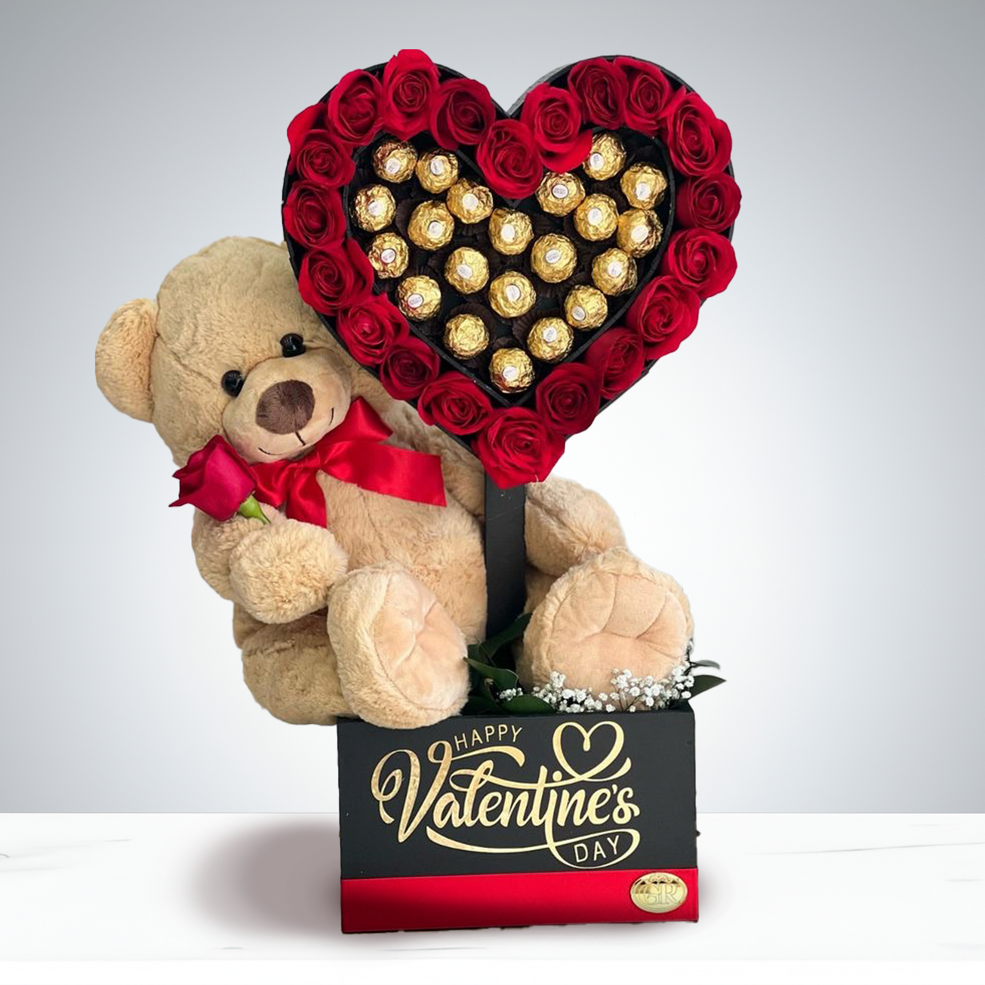 Luxury Heart by GIA - A romantic and elegant gift that combines sweetness and charm. This arrangement features a plush teddy bear holding a delicate red rose, seated beside a heart-shaped display filled with chocolates and surrounded by vibrant artificial roses. Presented in a sleek black box with golden lettering, it’s a perfect blend of luxury and love—ideal for expressing heartfelt emotions on special occasions