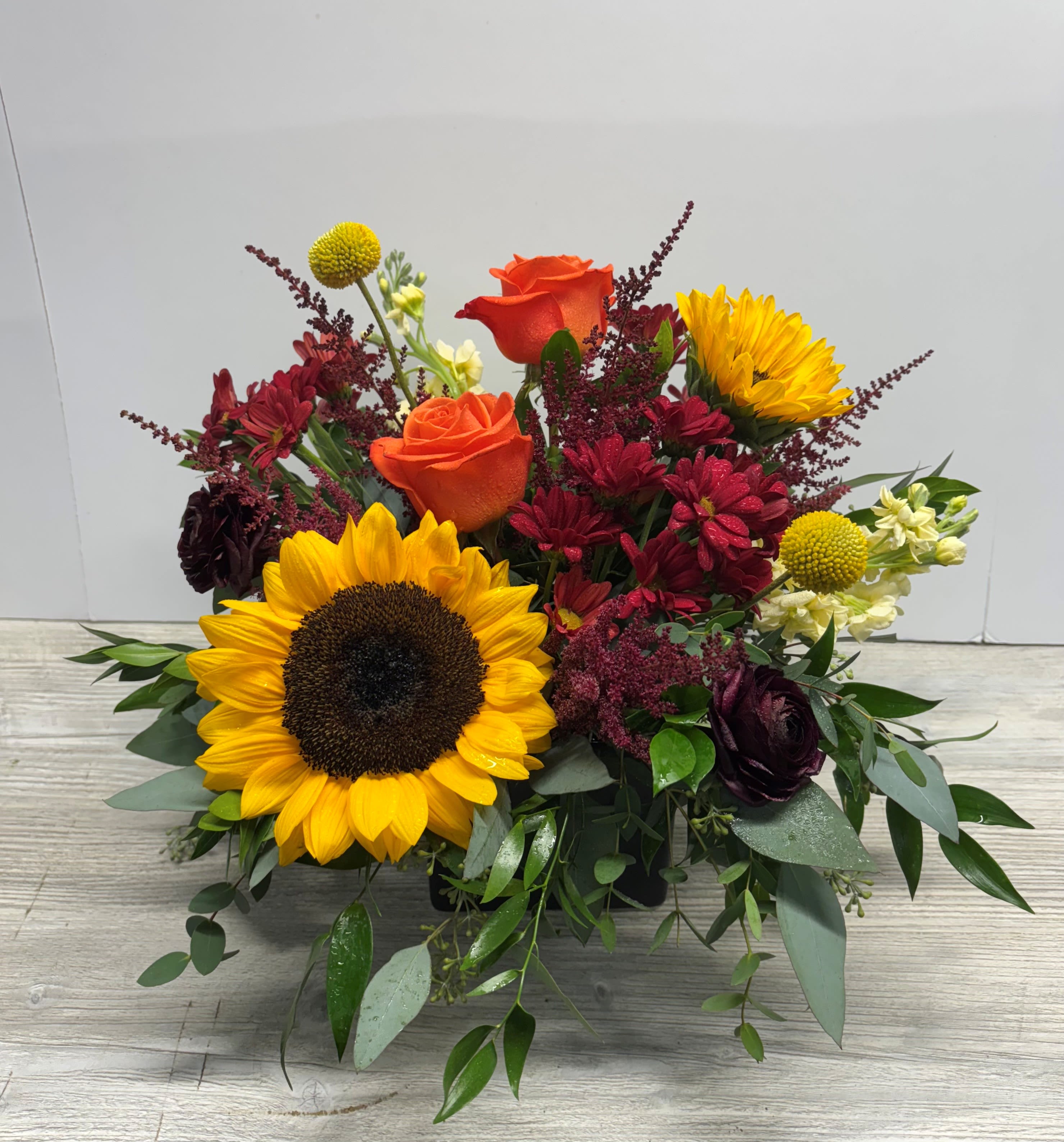 Low arrangement with a large sunflower, orange roses, red mums, and yellow accents in a black rectangular vase.