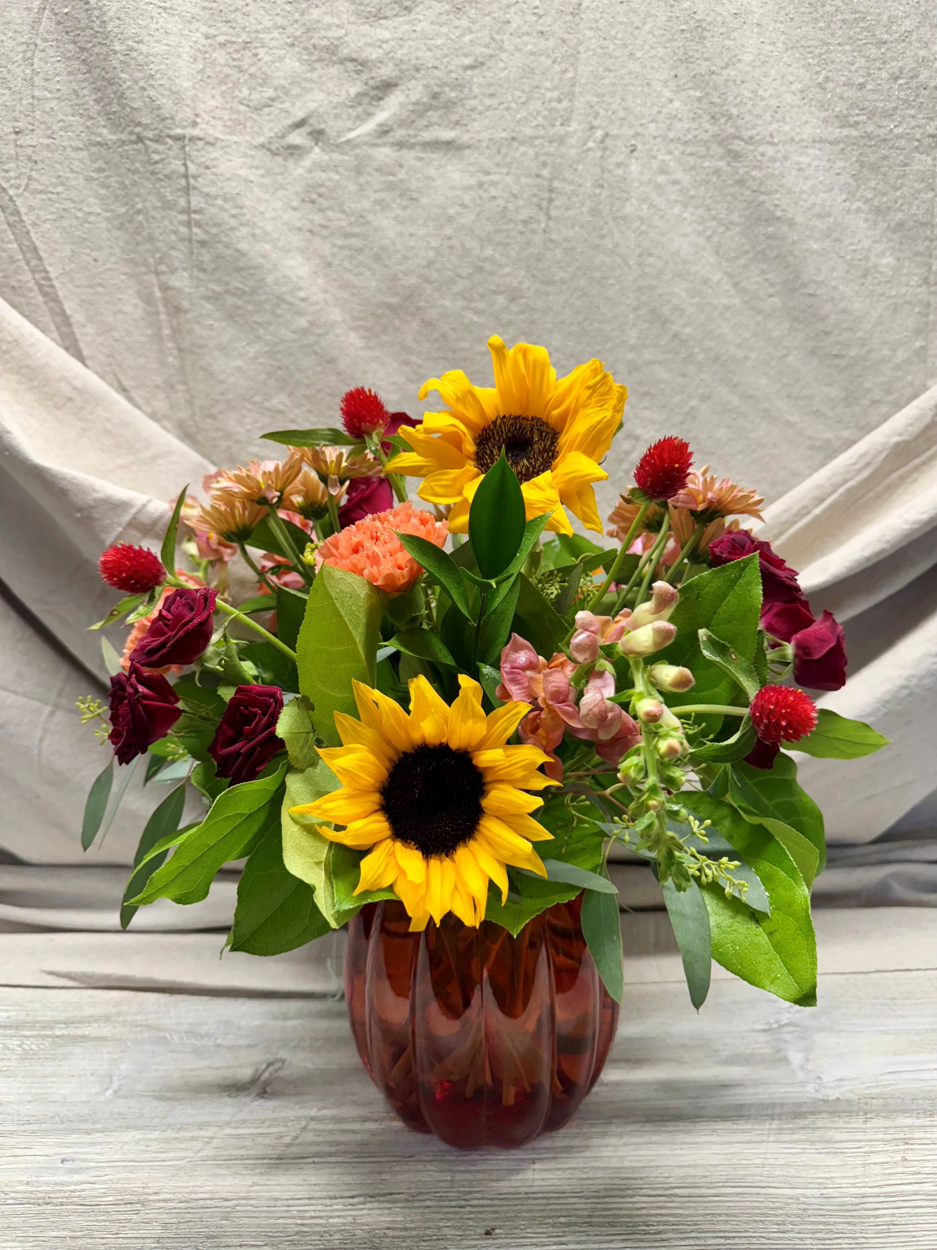 Mixed bouquet with sunflowers, red roses, and peach blooms arranged in a ribbed amber glass vase