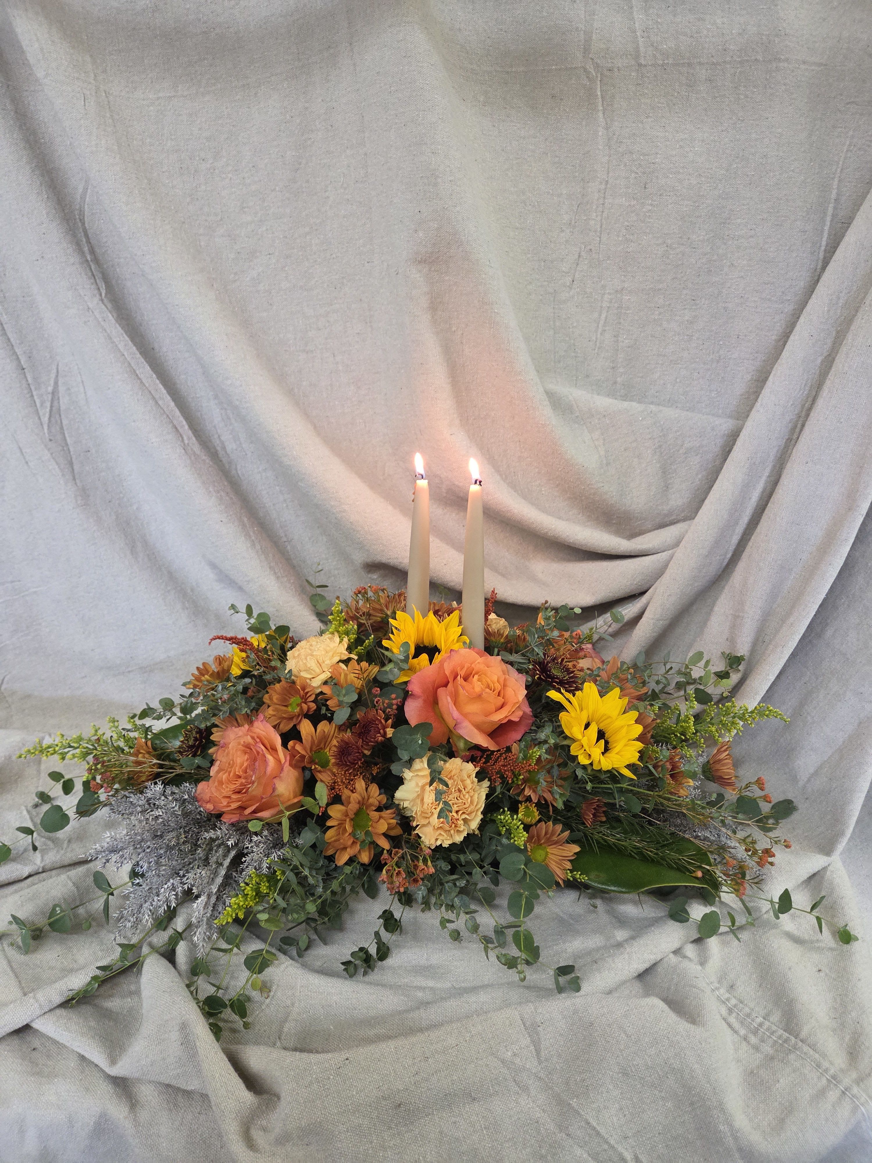Low autumn centerpiece of orange roses, yellow sunflowers, and two lit taper candles on a neutral cloth backdrop.