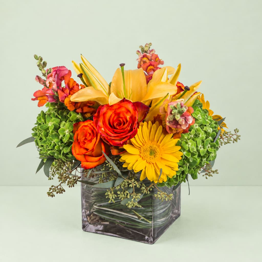 Bright mixed bouquet in a square glass vase with roses, lilies, and a yellow gerbera daisy