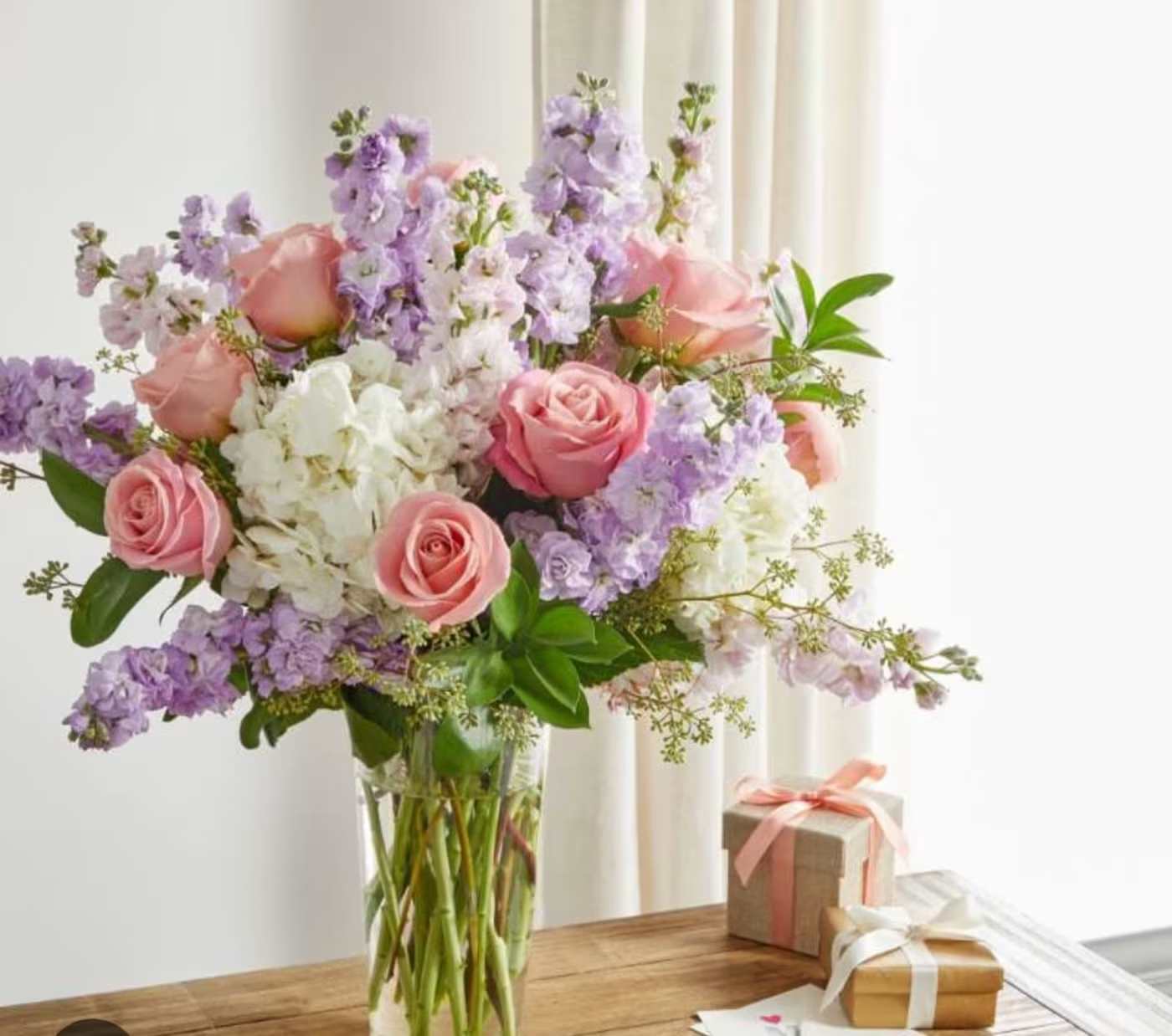 Pink roses and lavender flowers in a clear glass vase with gift boxes nearby
