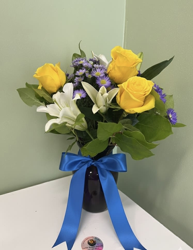 Arrangement of yellow roses, white lilies, and purple asters in a dark vase with a blue ribbon bow