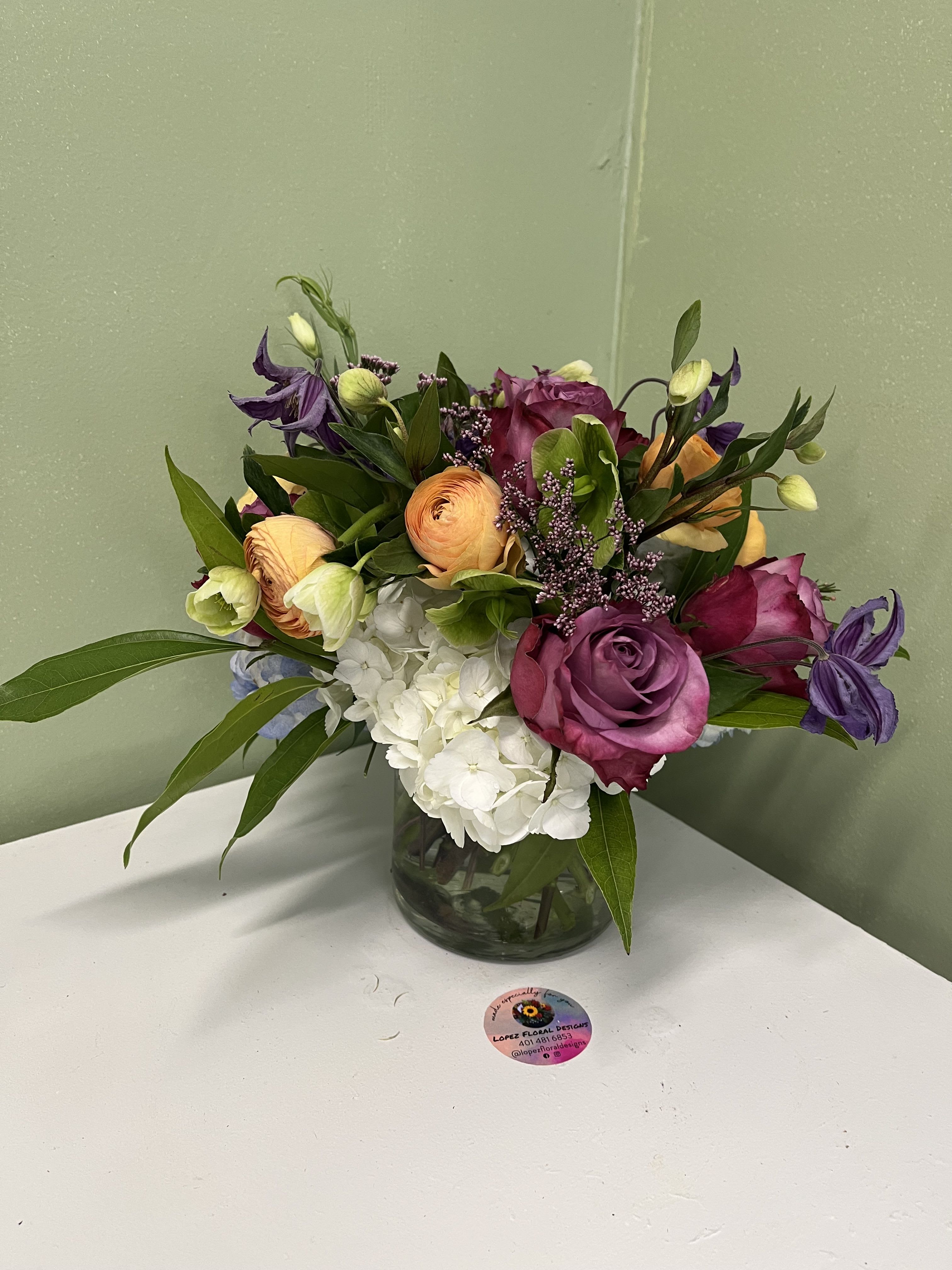 Low arrangement of purple roses, orange ranunculus, and white hydrangeas in a clear glass vase
