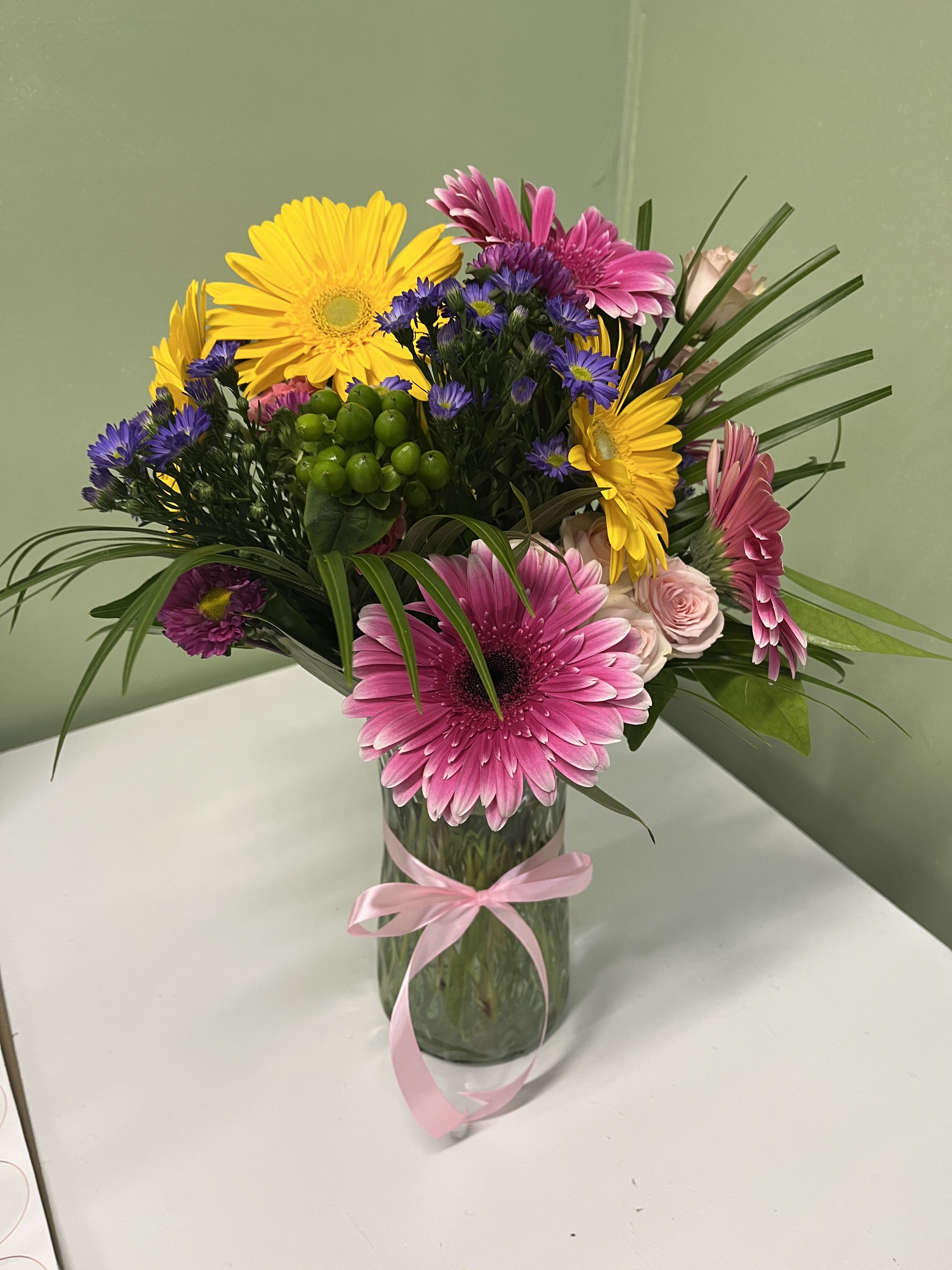 Tall vase arrangement of pink gerbera daisies, yellow gerbera daisies, purple asters, and pink roses.