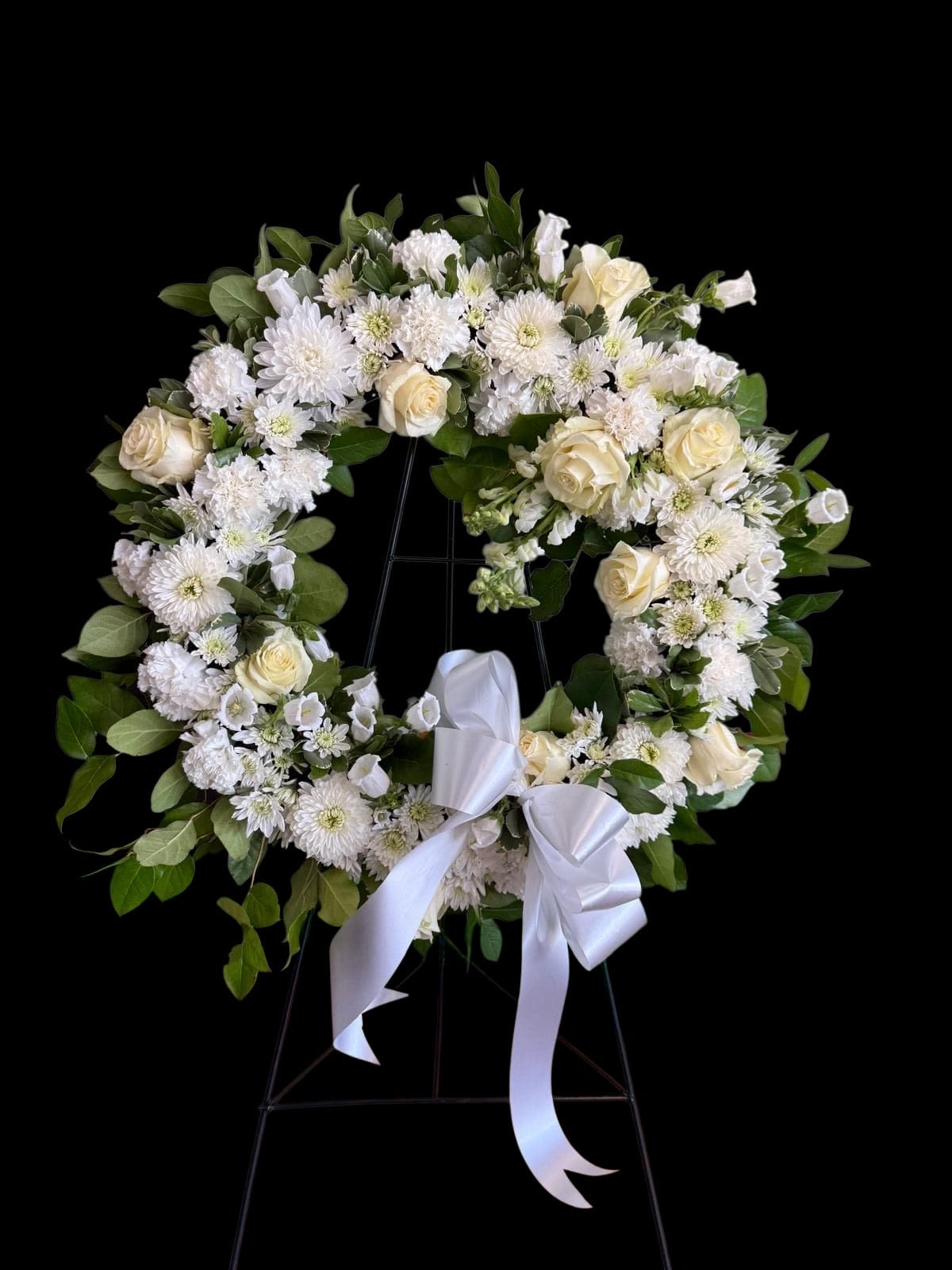 White floral wreath with roses and daisies on a stand, tied with a white ribbon