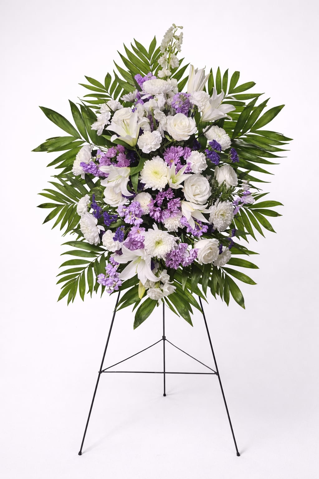 Standing spray of white lilies, roses, and purple mums with large palm leaves on a wire easel.