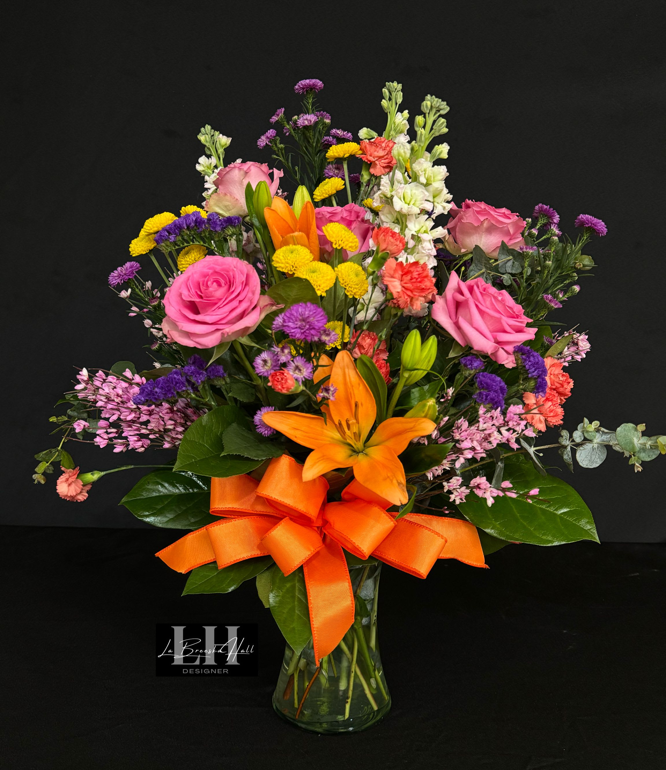 Mixed bouquet of pink, orange, yellow, and purple flowers in a glass vase with an orange ribbon