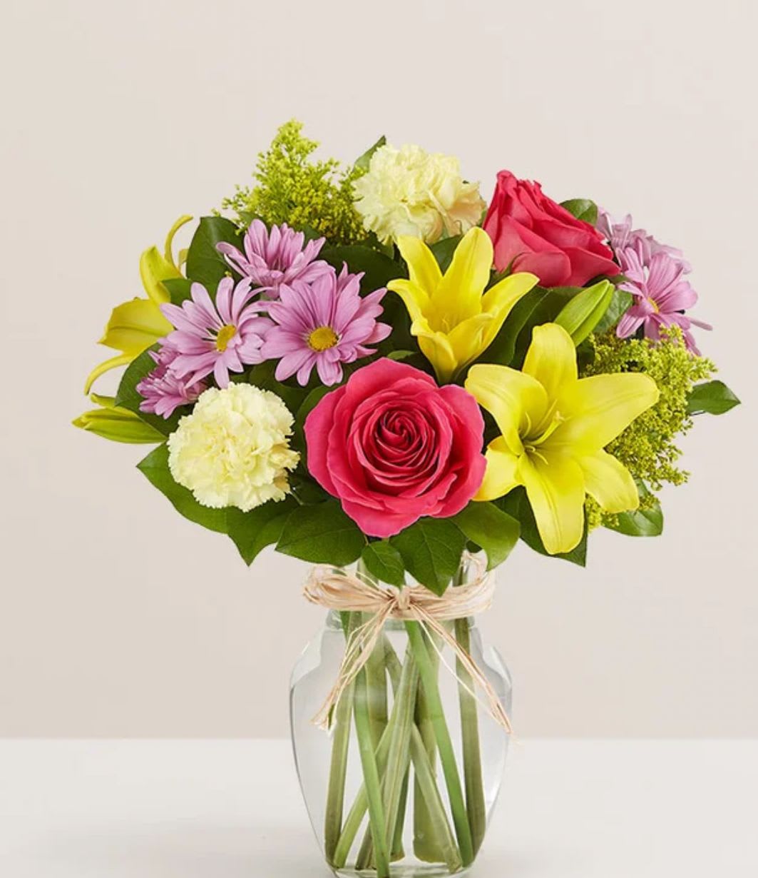 Fields of Europe for Mom - All-around arrangement with hot pink roses; yellow Asiatic lilies and carnations; lavender daisy poms; solidago; accented with assorted greenery Artistically designed in clear glass vase tied with raffia; measures 8"H Premium arrangement measures approximately 18"H x 14"W Deluxe arrangement measures approximately 17"H x 13"W Standard arrangement measures approximately 16"H x 12"W Our florists hand-design each arrangement, so colors, varieties and container may vary due to local availability To ensure lasting beauty, Asiatic lilies may arrive in bud form and will fully bloom over the next few days