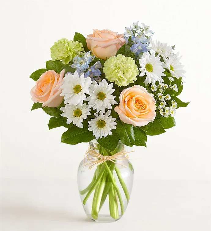 Summer Dunes Bouquet  - All-around arrangement with peach roses, blue delphiniums, lime-green carnations, and white daisy poms and monte casino, accented with assorted greenery Artistically designed in a clear glass vase accented with hand-tied raffia; measures 8"H Premium arrangement measures 19"H x 15"W Deluxe arrangement measures approximately 18"H x 14"W Standard arrangement measures approximately 17"H x 13"W