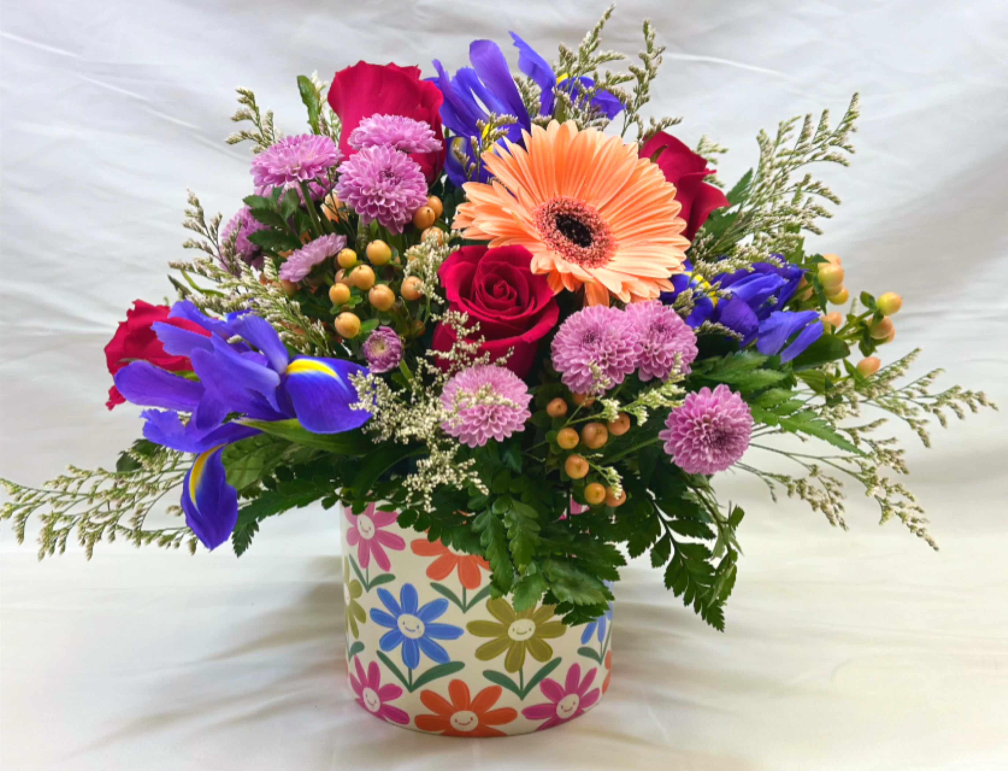 Give a Smile - The Leary's Flowers exclusive design is sure to live up to its name! The cheerful  gerbera daisies, along with iris and rose blossoms, are sure to make anyone smile! The vase is just as whimsical with colorful smiley faced flowers.  If the featured blooms are not available we will design your arrangement using the freshest flowers of equal or greater value.