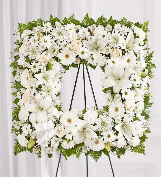 White floral wreath on a black easel with lilies, daisies, and roses