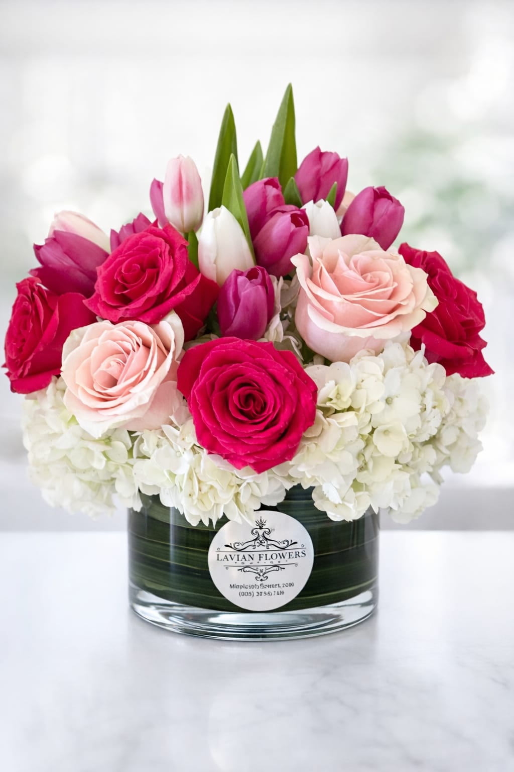 Pink Beauty  - A luxurious blend of hot pink and blush roses, fresh tulips, and soft white hydrangeas, this elegant floral arrangement is designed to make a lasting impression. Arranged in a modern glass vase with lush greenery, this piece brings together vibrant color 