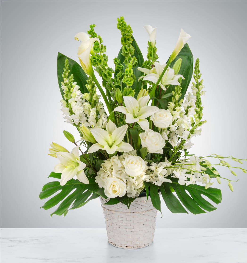 White lilies and roses arranged in a white basket with broad green leaves.