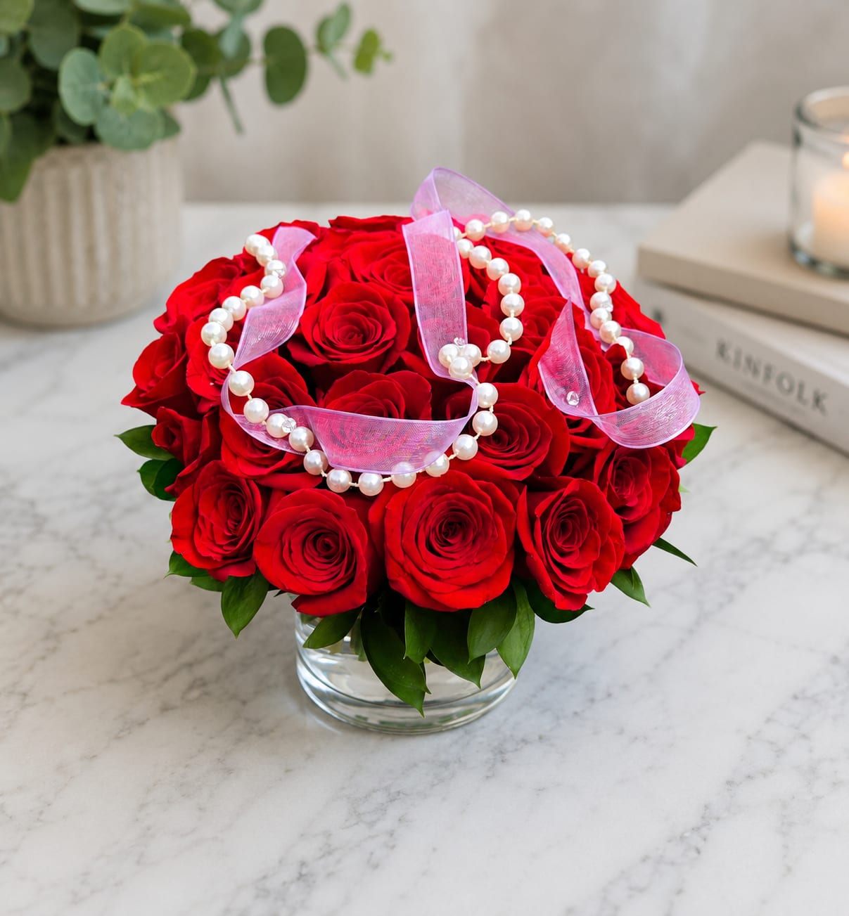 Red roses arranged in a clear glass vase with pink ribbon and pearl strands