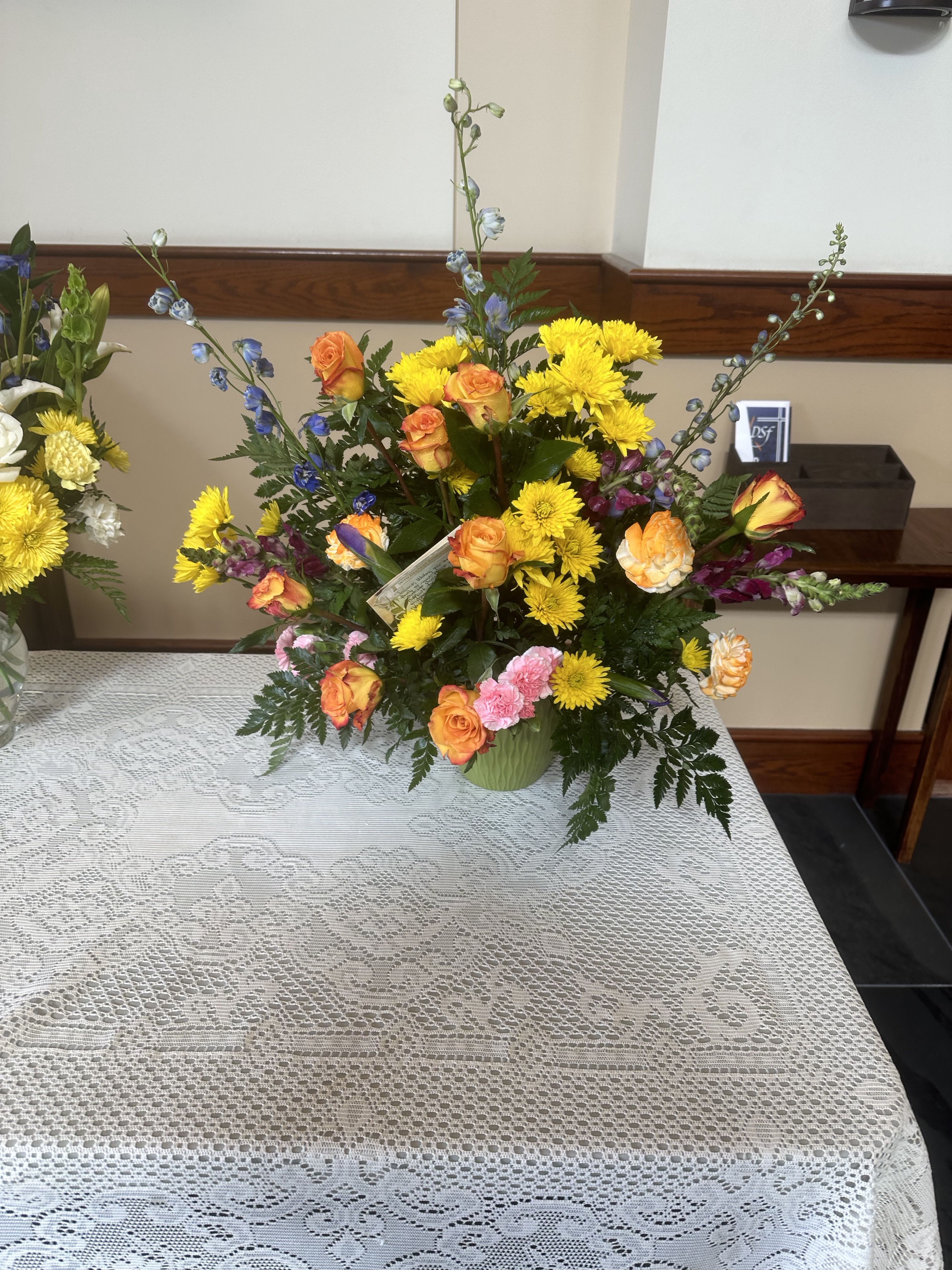 Tall mixed arrangement of yellow mums, orange roses, and blue delphinium in a green vase on a lace tablecloth