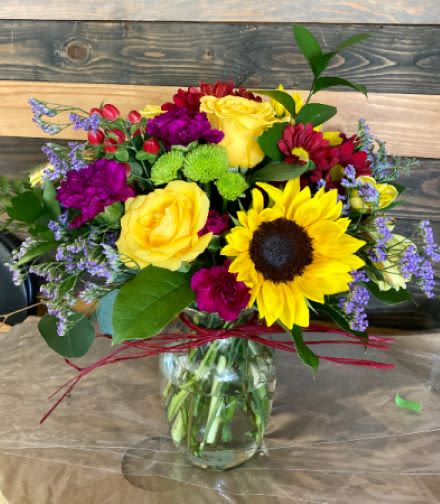 Mixed bouquet with yellow sunflower, yellow roses, and purple flowers in a clear glass jar