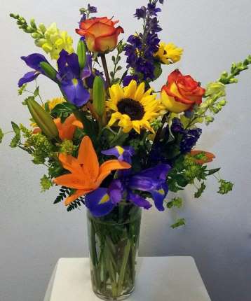 Tall mixed bouquet with orange lilies, yellow sunflowers, purple irises, and roses in a clear glass vase