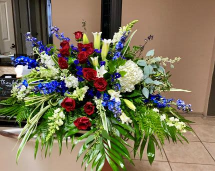 Large red, white, and blue floral spray with roses, lilies, and delphinium in an elongated design