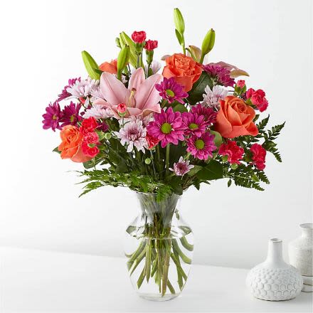 Mixed bouquet of pink lilies, orange roses, and bright daisies in a clear glass vase