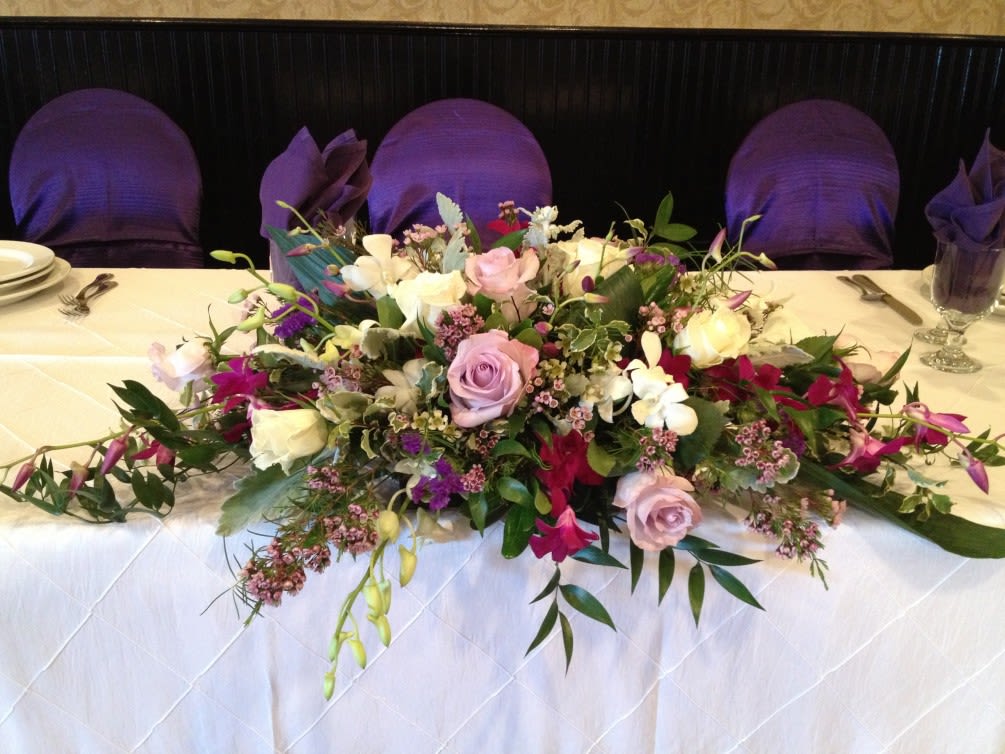 Low centerpiece of pink roses, white blooms, and purple flowers arranged across a banquet table