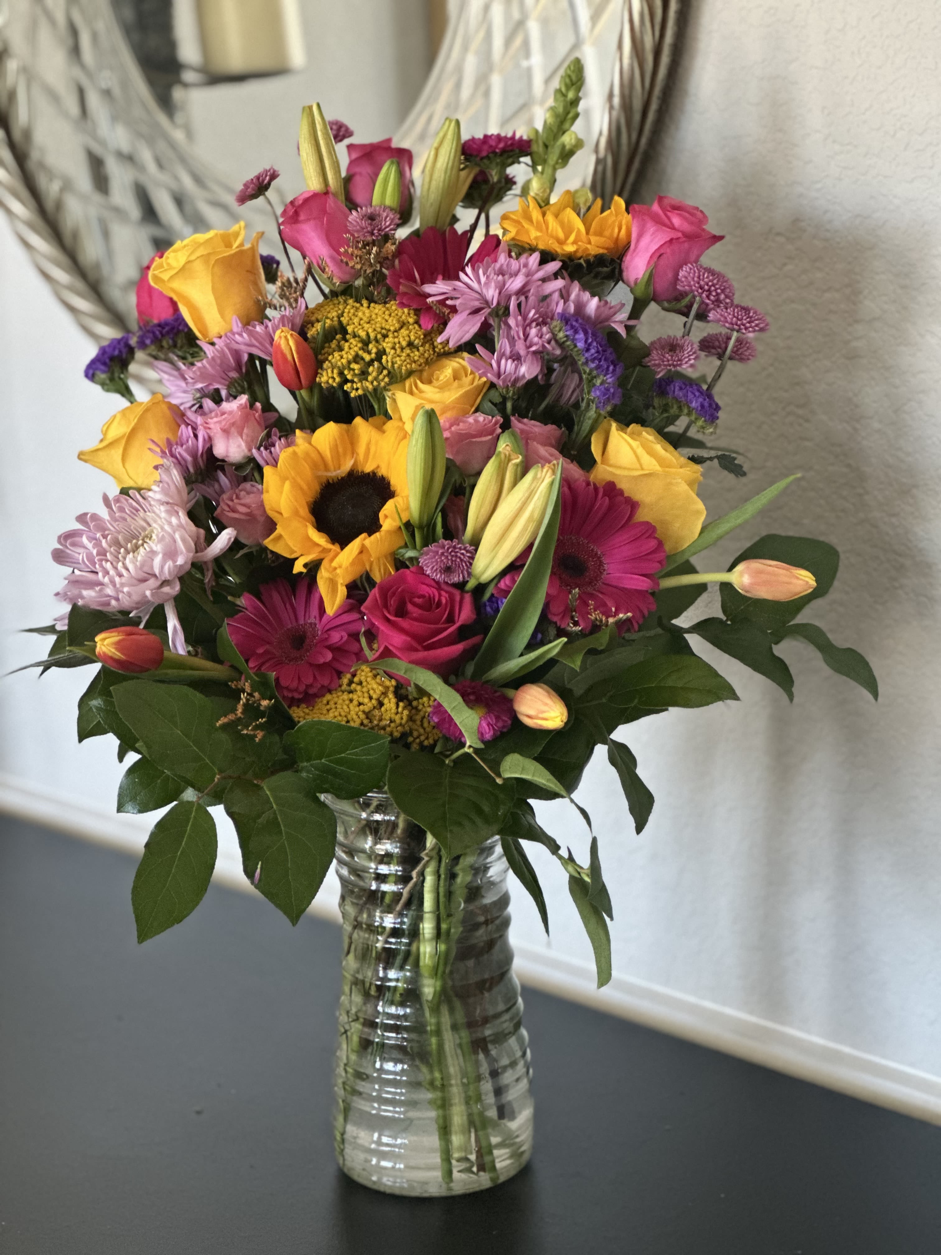 Tall mixed bouquet of sunflowers, roses, lilies, and other bright blooms in a clear glass vase