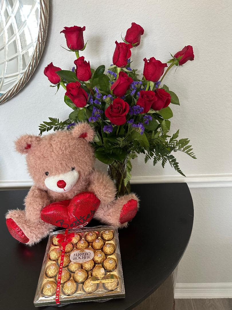 Red rose bouquet in a glass vase with purple blooms, a plush bear with red heart, and a box of chocolates.