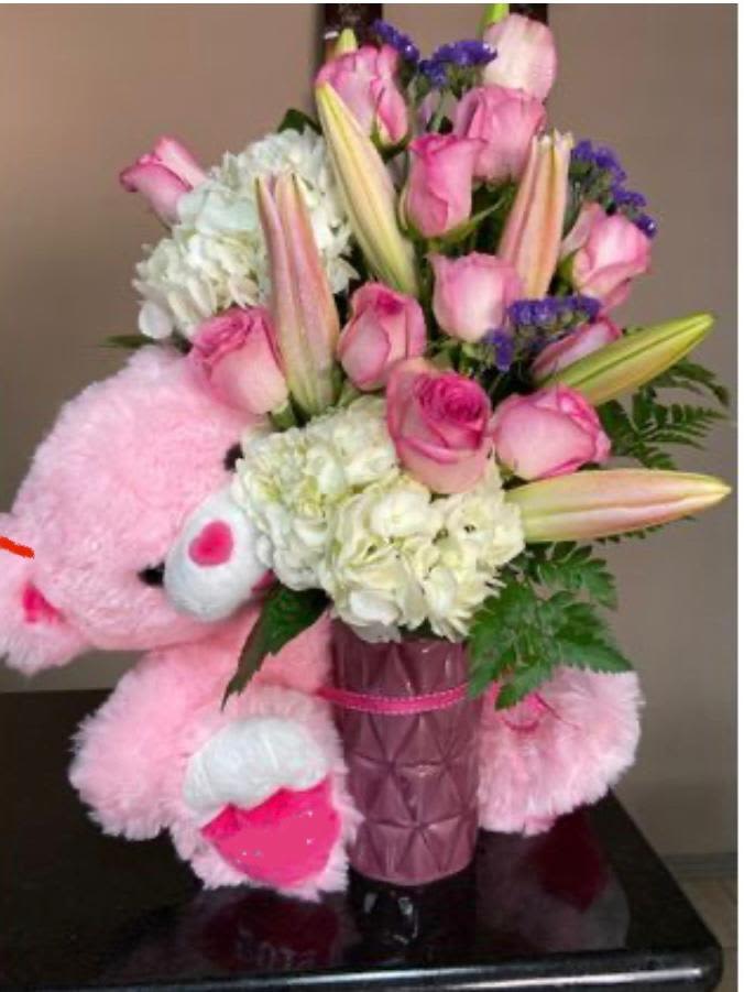 Pink teddy bear hugging a vase of pink roses, lilies, and white hydrangeas